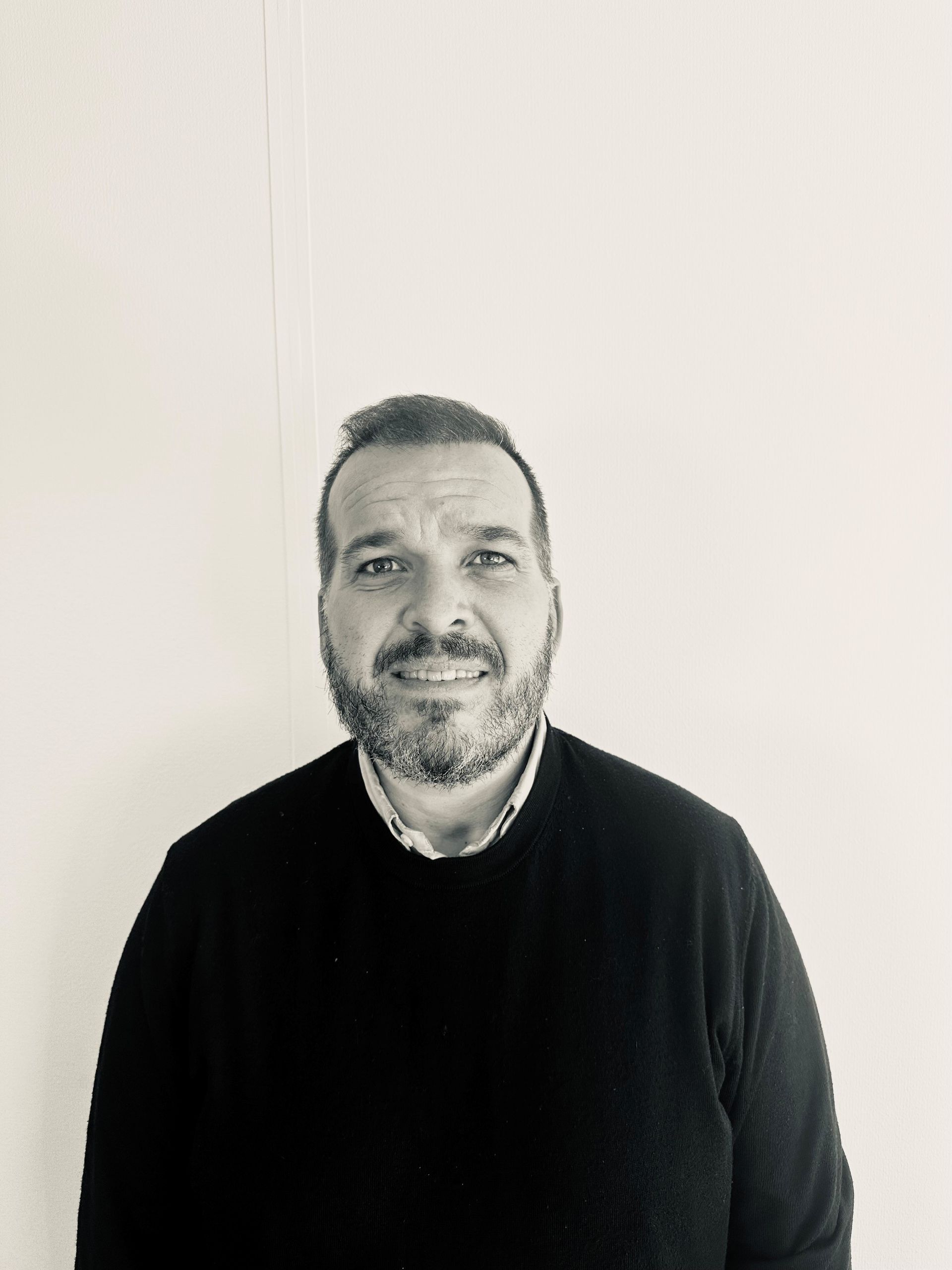 Smiling man in a black sweater against a plain light wall.