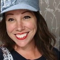 Woman smiling, wearing a denim train conductor's cap, red lipstick, and dark top.