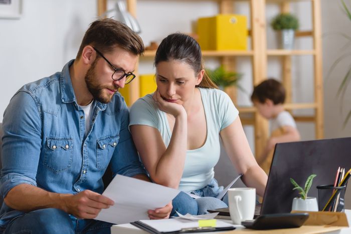 A couple reviews documents at a laptop, appearing stressed. A child plays in the background.