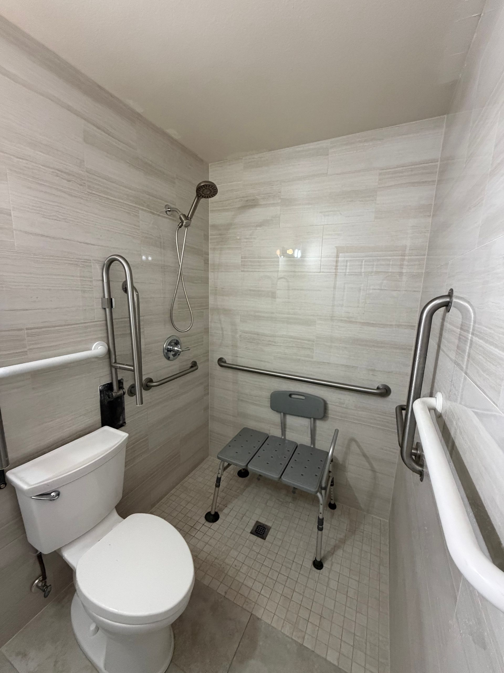 Accessible bathroom with shower, toilet, grab bars, and shower chair. Beige tile and neutral tones.
