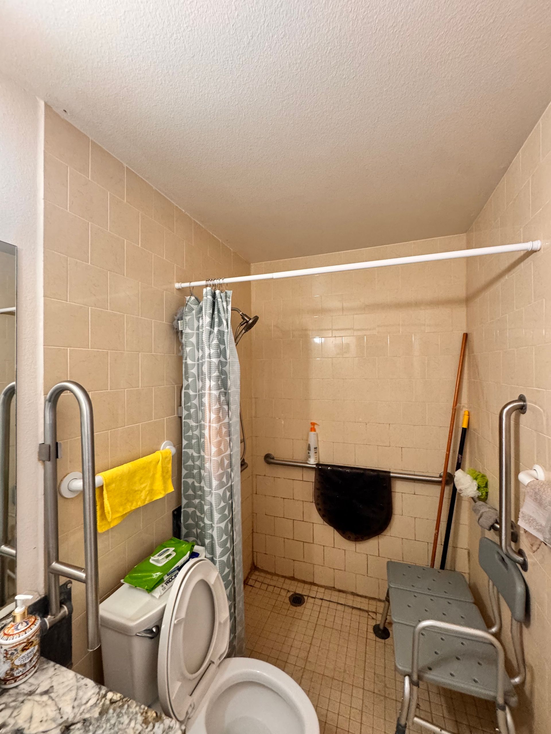 Accessible bathroom with shower, toilet, grab bars, and shower seat. Beige tile, curtain, and towel present.