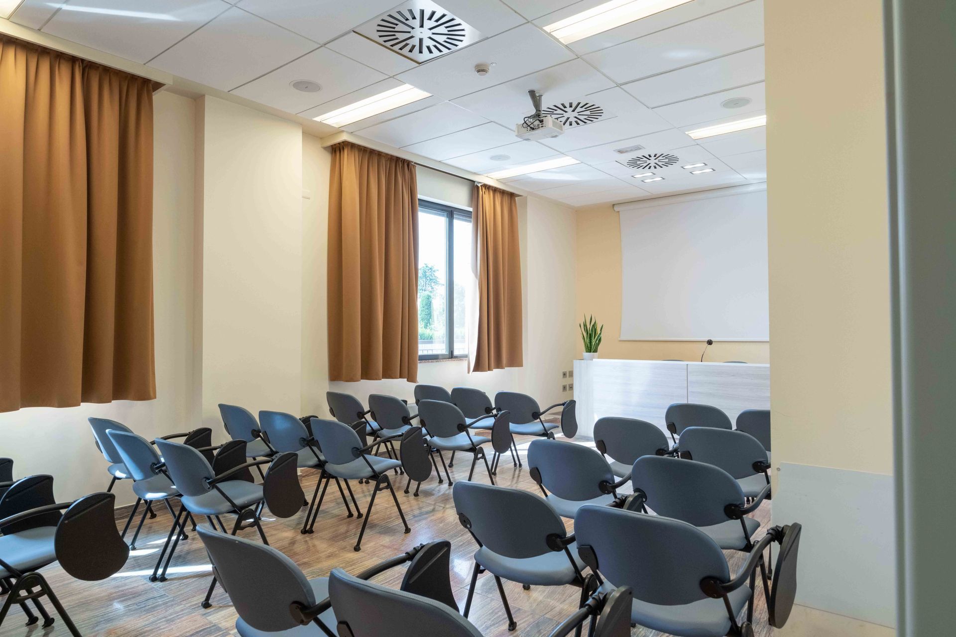 Aula vuota con file di sedie di fronte a uno schermo per proiettore, tende e un podio.