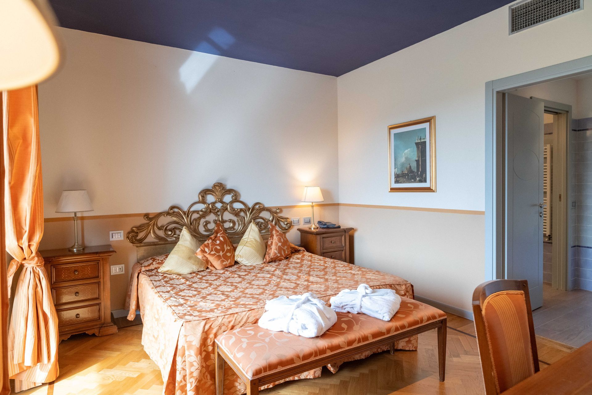 Camera d'albergo con letto, panca, comodini e porta per il bagno.