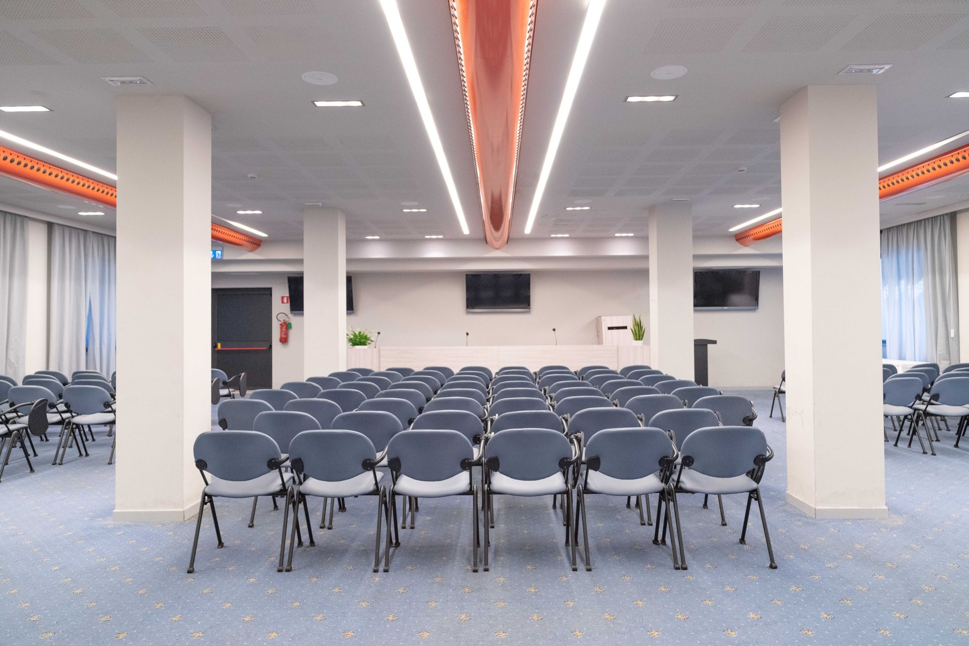 File di sedie blu sono disposte di fronte a un palco in una sala conferenze con pilastri bianchi e moquette blu.