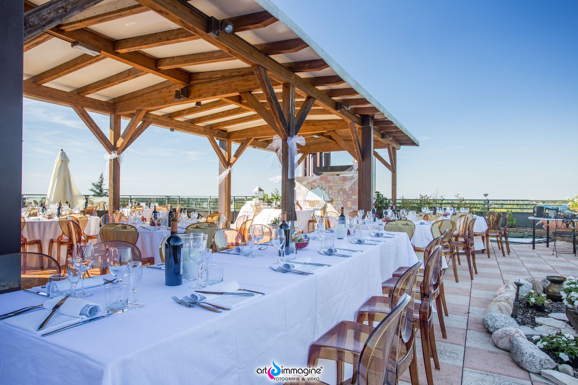 Ricevimento di nozze all'aperto con lungo tavolo, tovaglia bianca e baldacchino in legno sotto un cielo azzurro e terso.