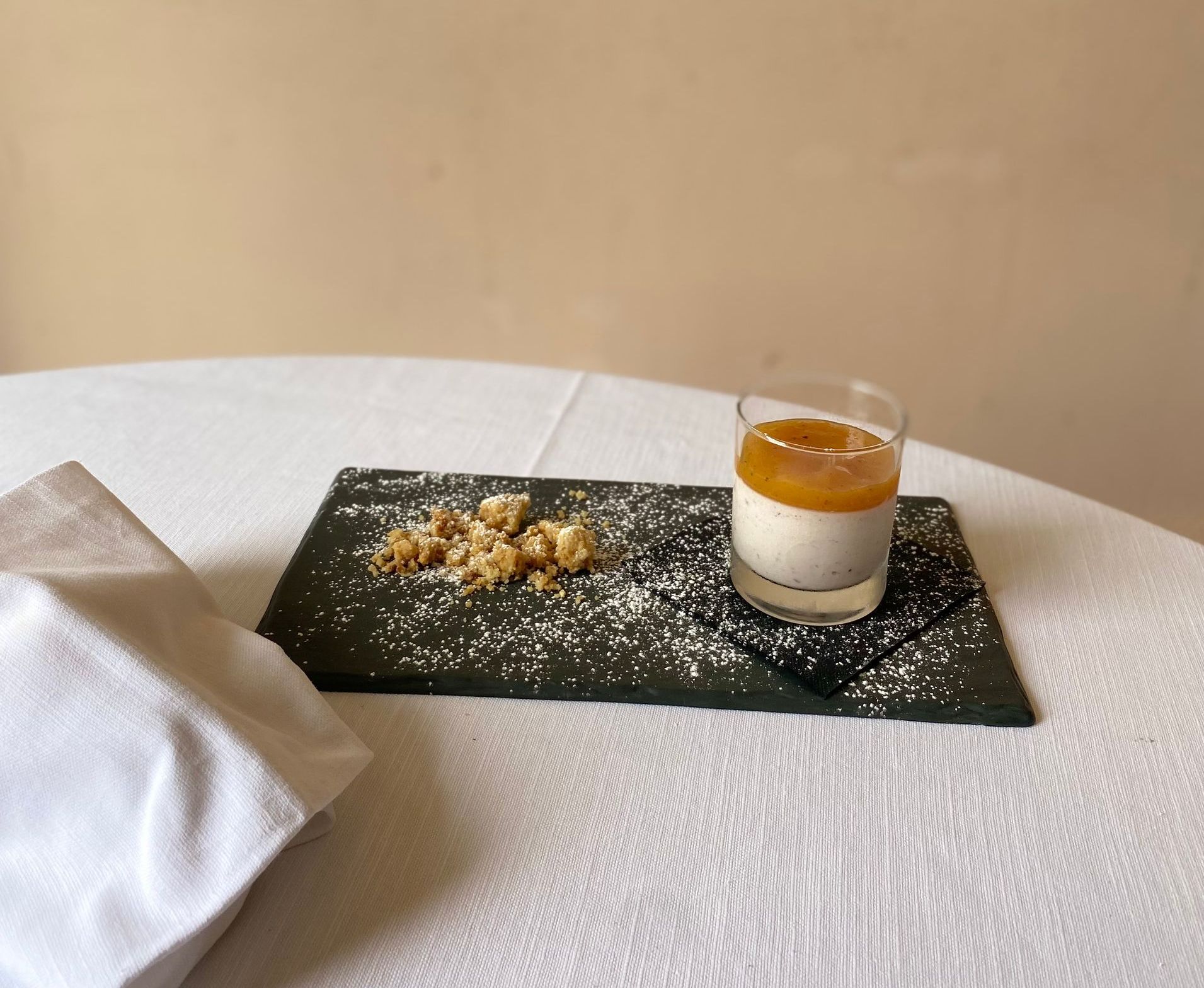 Dessert su una lavagna nera: strati di crema pasticcera, crumble e un bicchiere, il tutto su una tovaglia bianca.