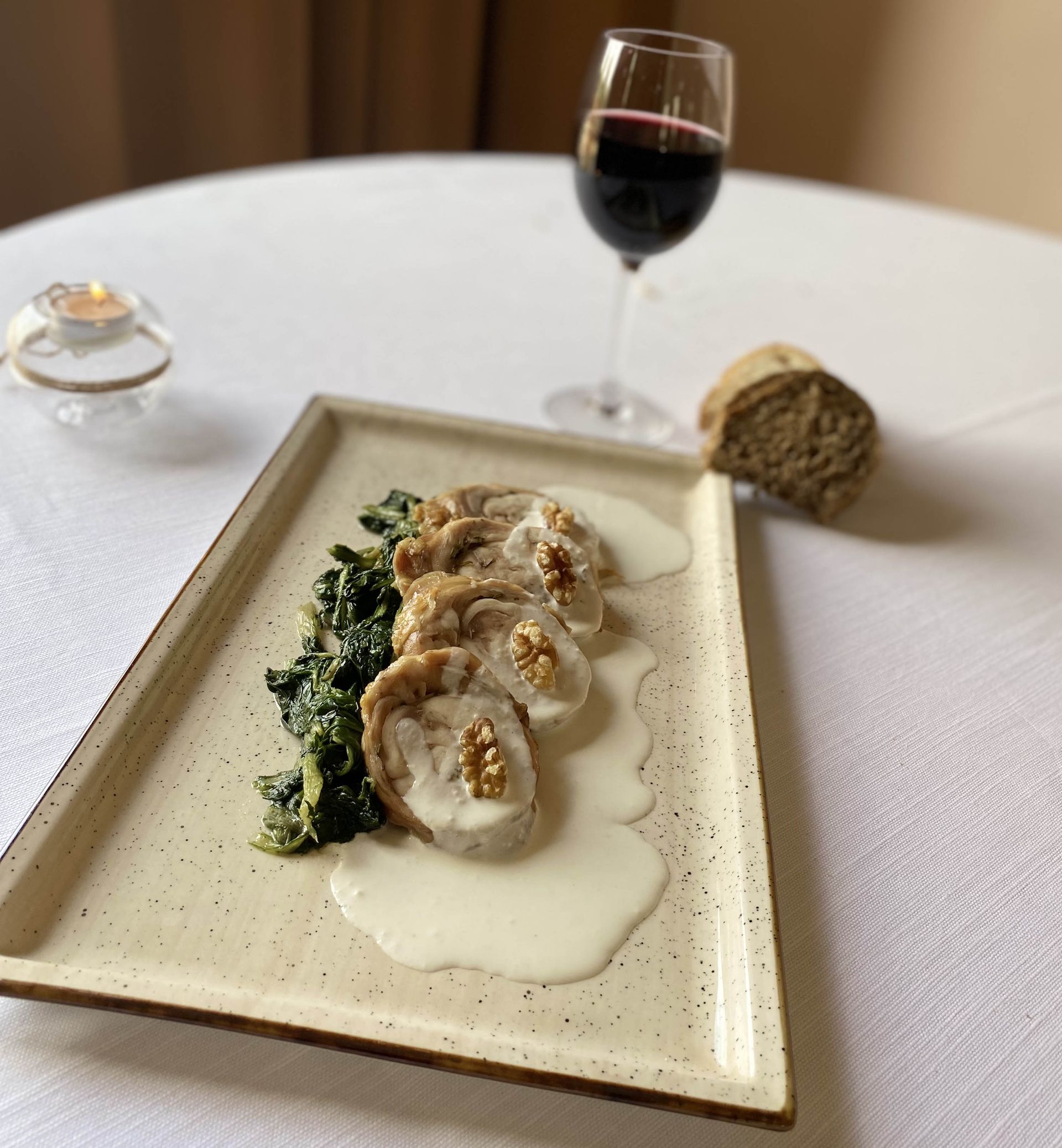 Piatto con carne affettata, spinaci, salsa cremosa, noci, pane, vino. Tavola apparecchiata con molta luce.