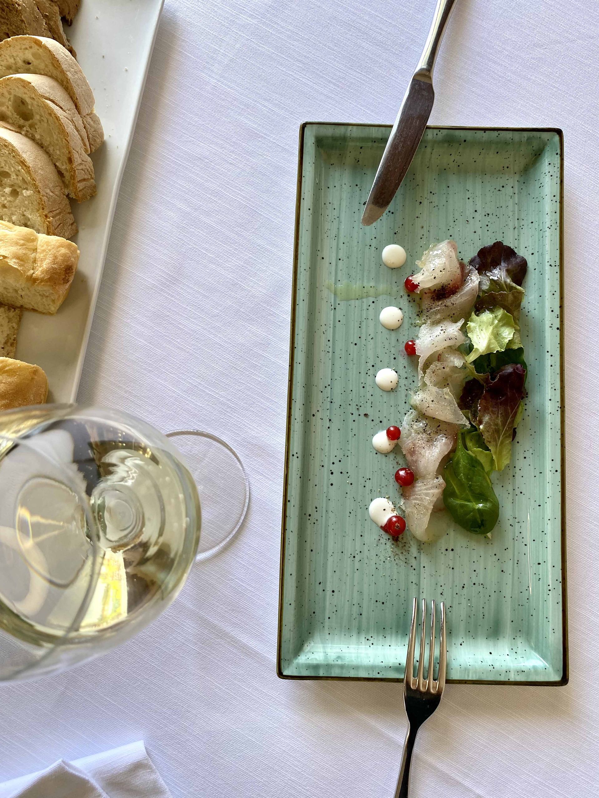 Piatto di pesce affettato con salsa e verdure, pane e teiera su una tovaglia bianca.