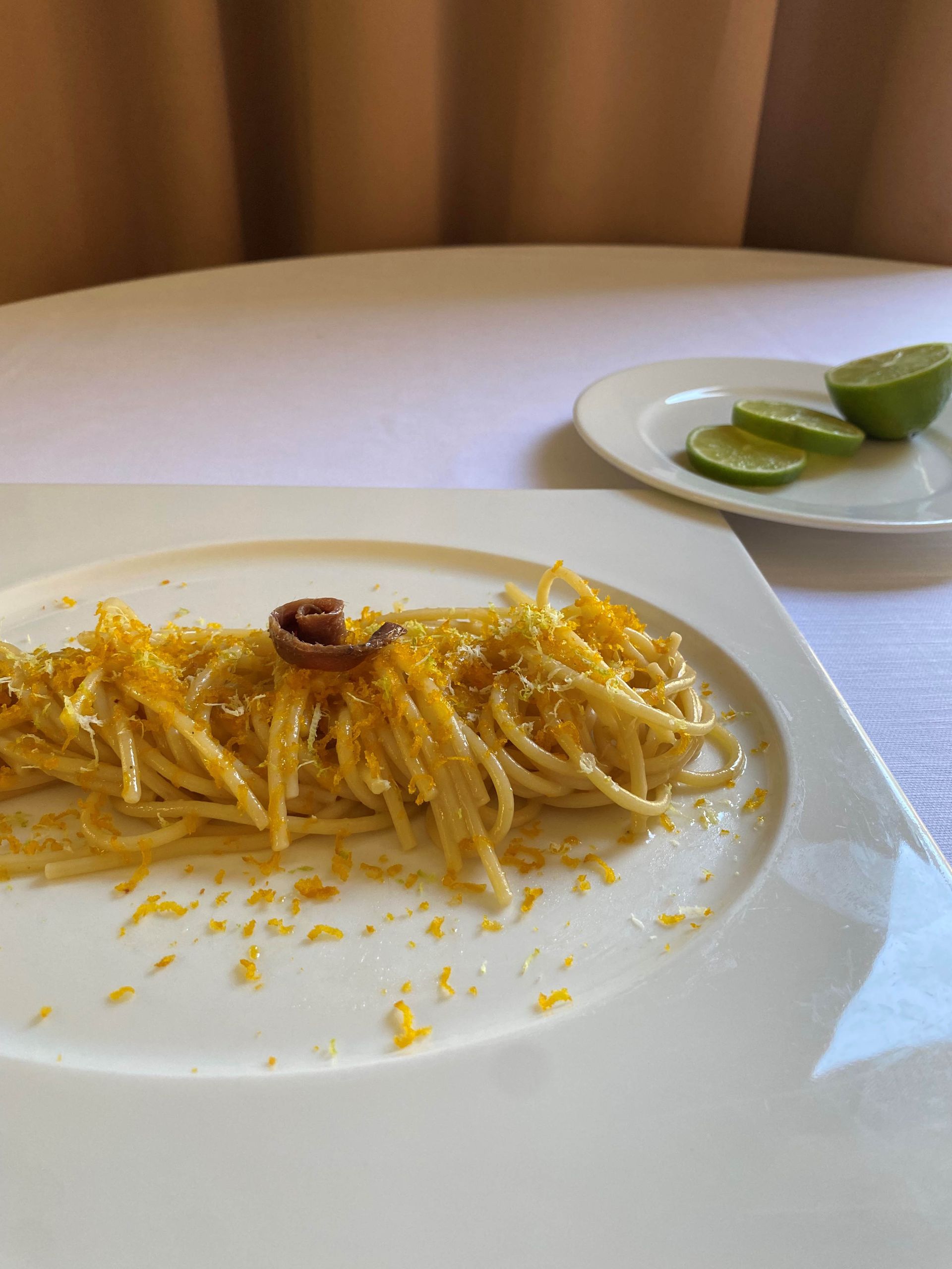 Piatto di pasta su un piatto bianco, guarnito con scorza gialla e un piccolo condimento marrone. Lime a fette su un piattino.