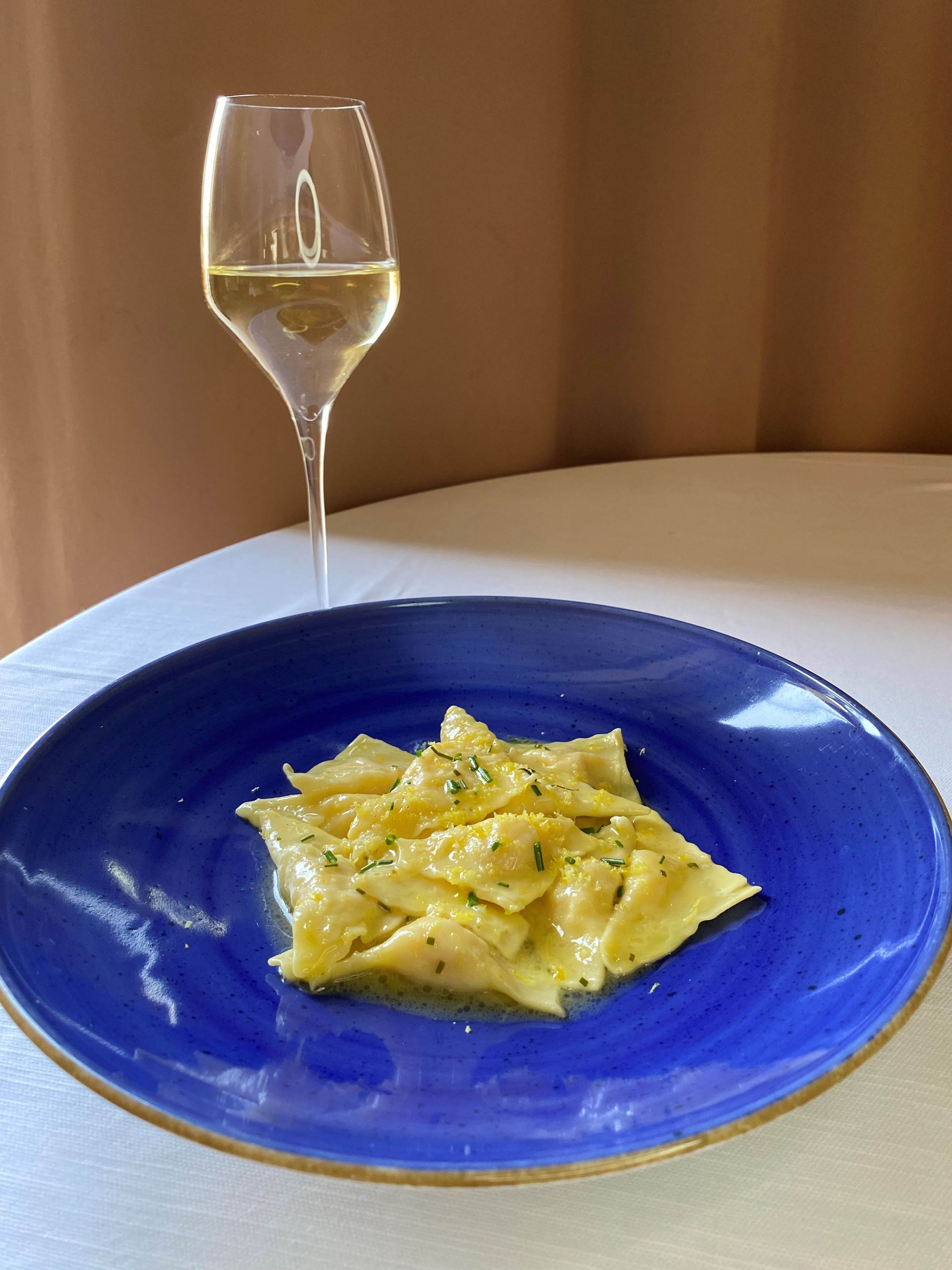 Piatto di pasta blu con un bicchiere di vino sullo sfondo. La pasta è gialla. Il vino è bianco.