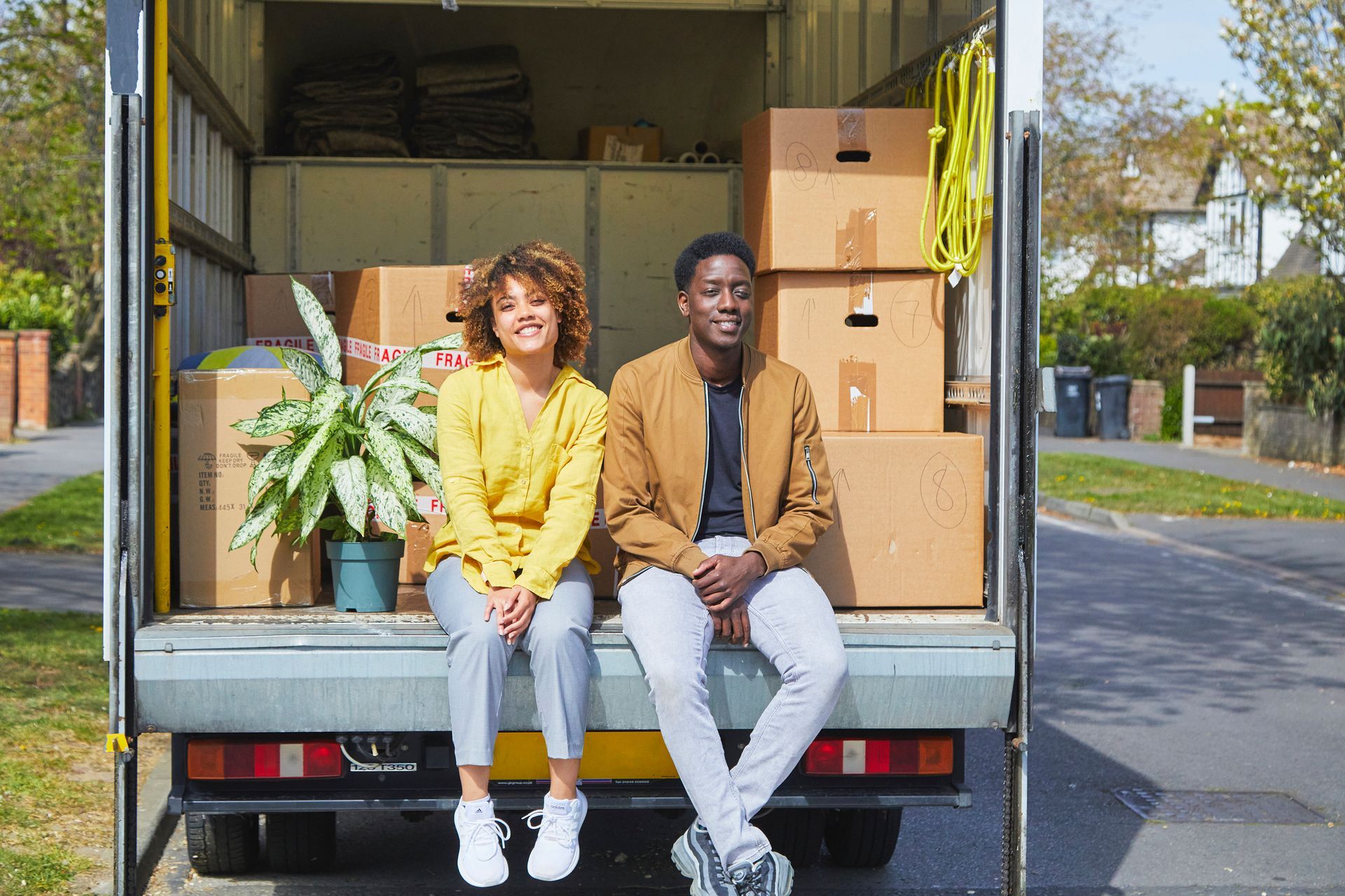 People are sitting on a moving truck loaded with boxes and a plant.