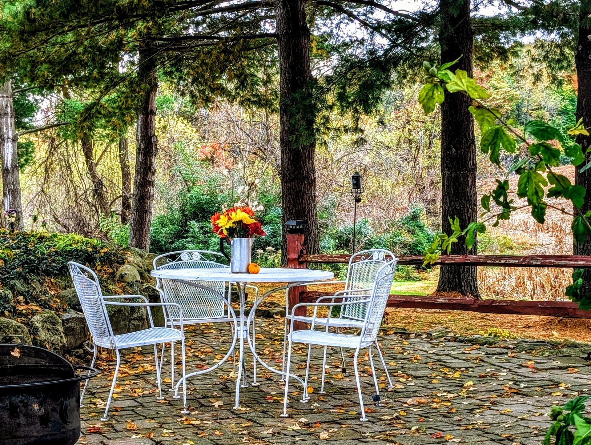 White patio set with flowers on a table in a wooded outdoor area.
