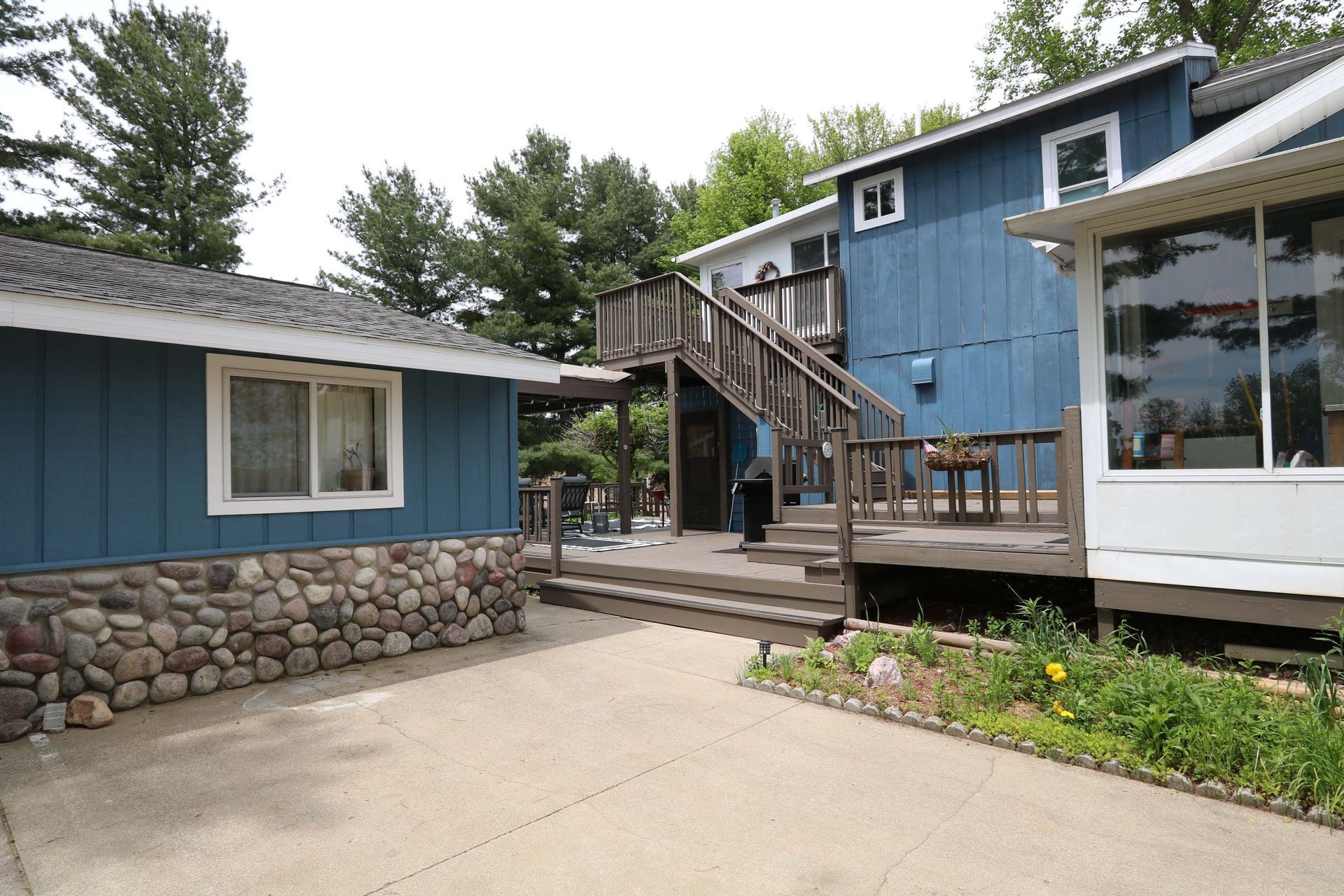 Blue house with wooden deck, stone foundation, and driveway. Green plants and trees in the background.