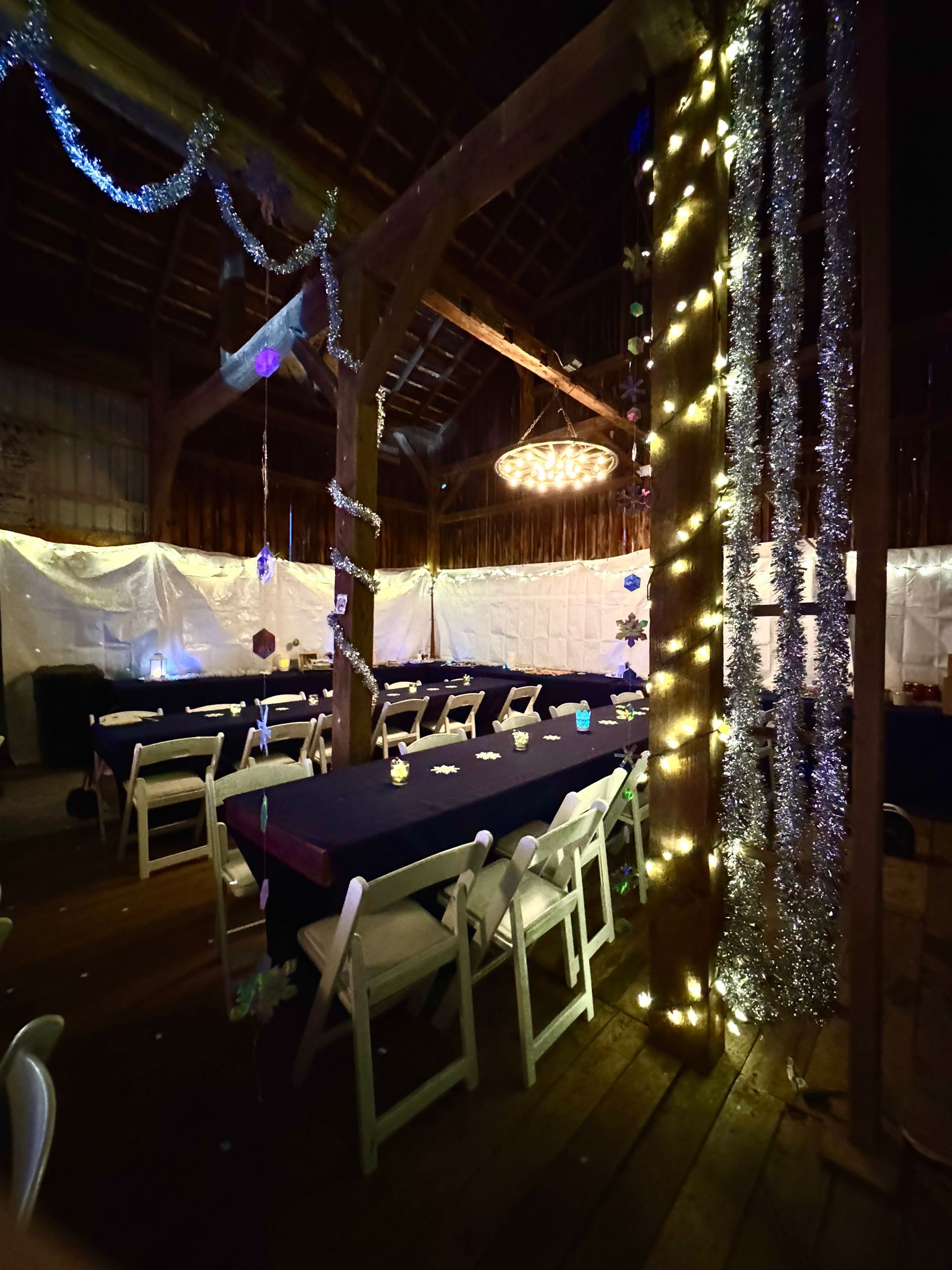 Barn decorated for an event, long tables with blue tablecloths, white chairs, and string lights.