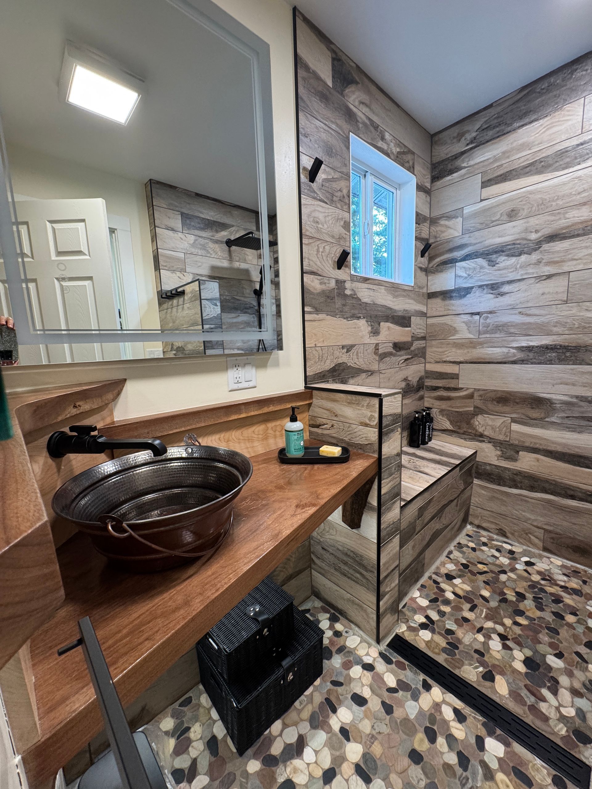 Bathroom with wood-look tile, copper sink, and pebble floor. A large mirror reflects a white door.