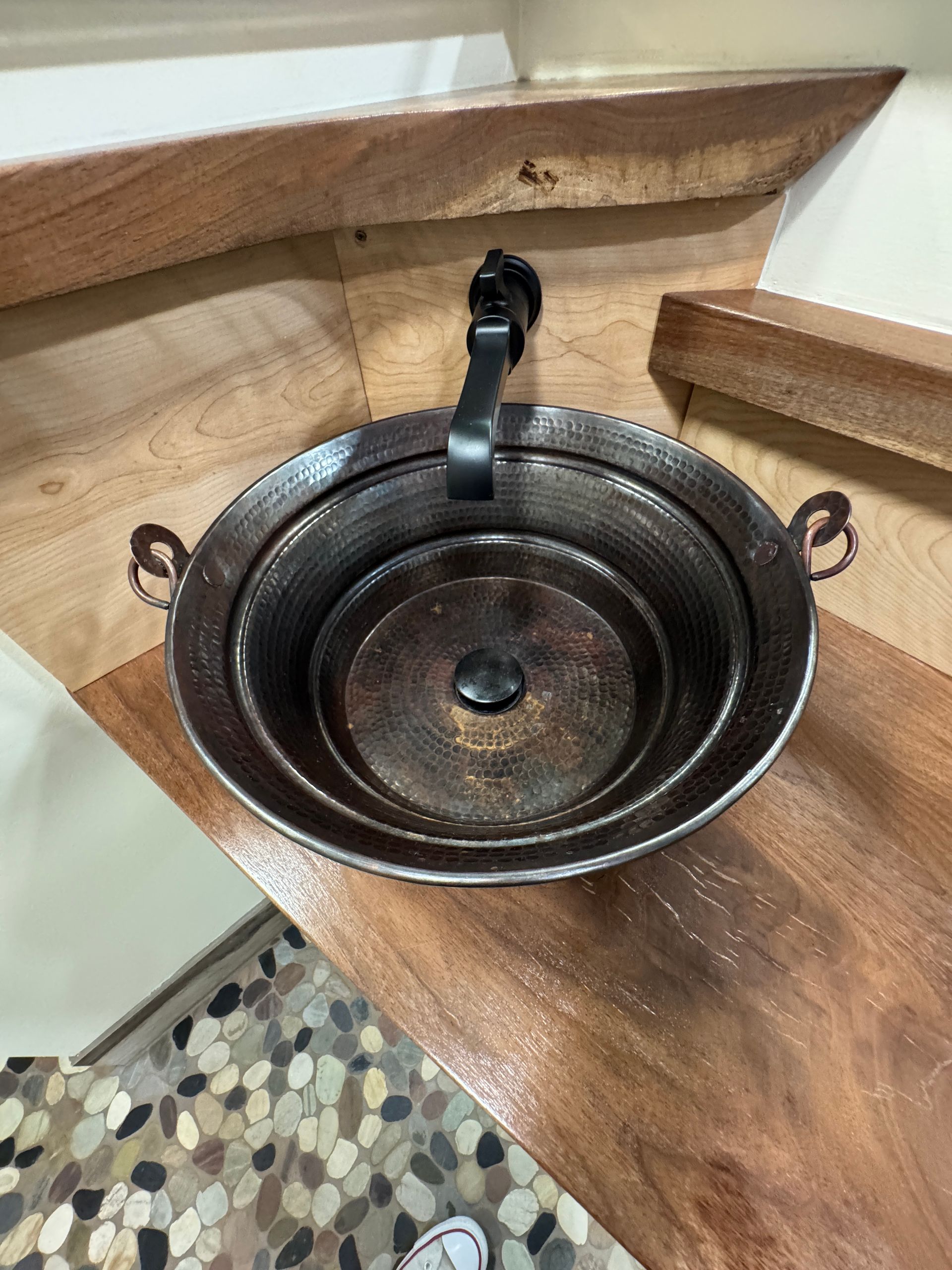 A round, dark  copper bucket sink with black a faucet, set into a wooden countertop.