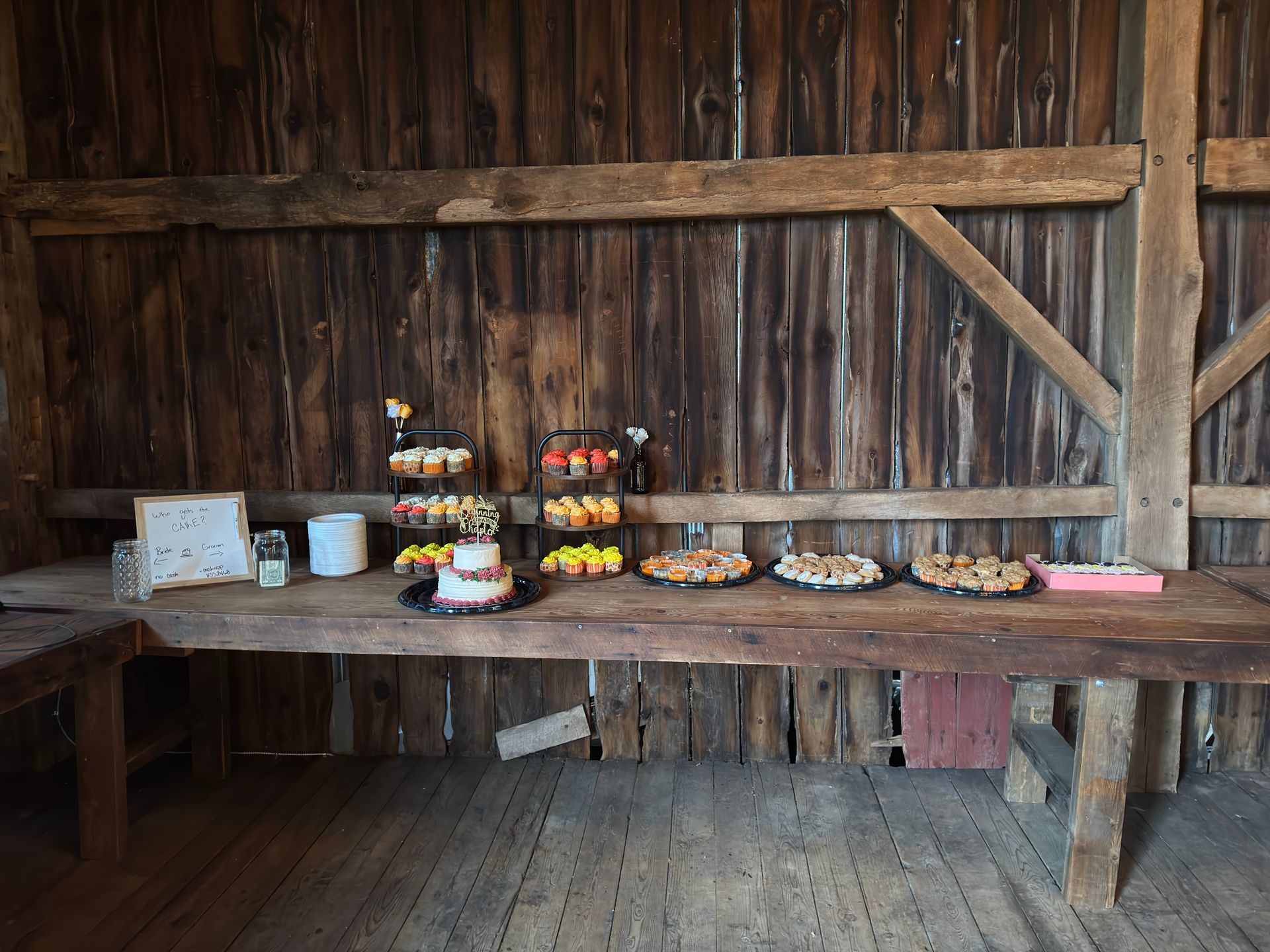 Wooden table laden with cupcakes, cake, and pastries inside a rustic wedding and event venue barn.