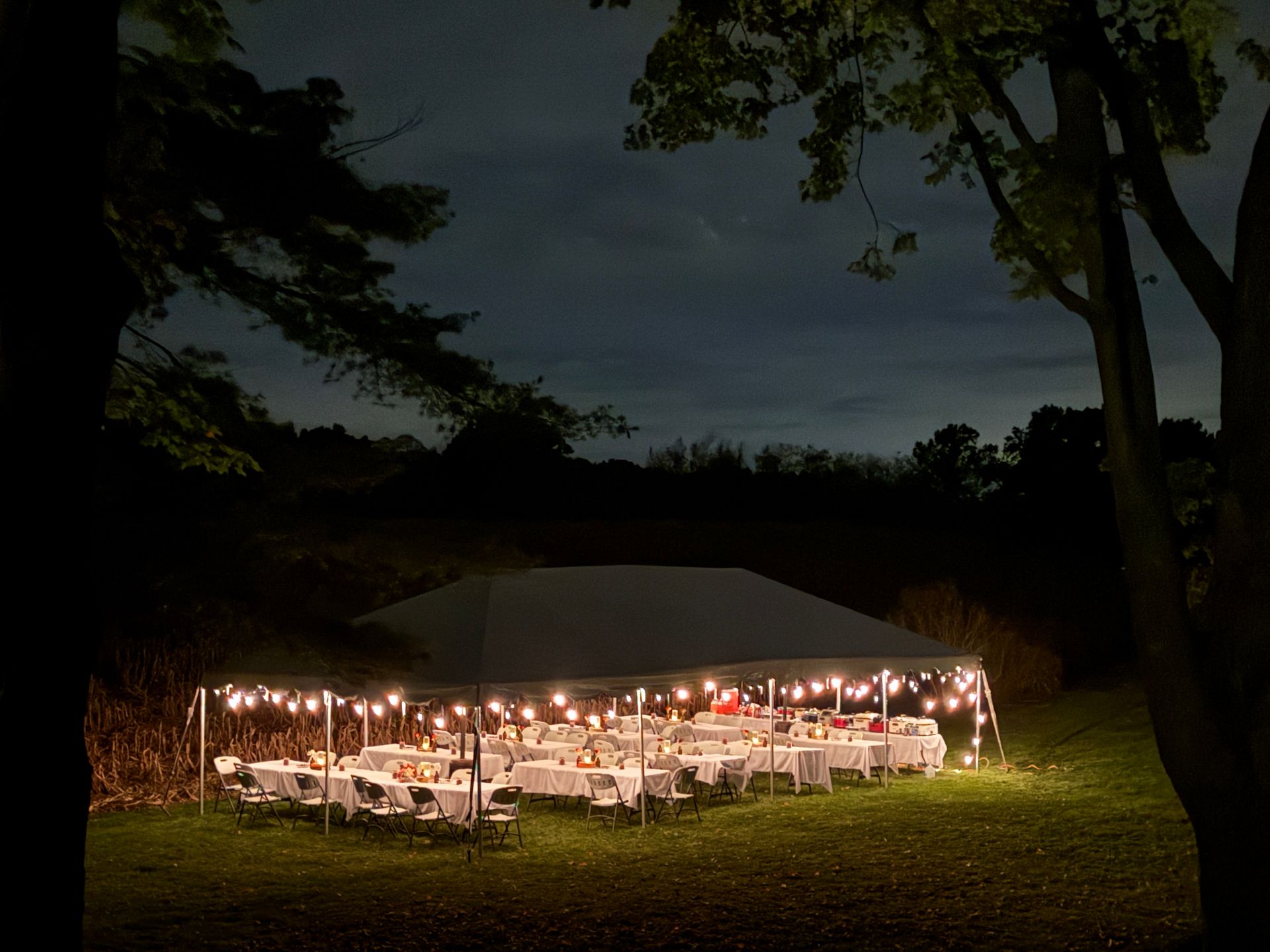 Lit tent with tables and string lights on a grassy lawn at night.