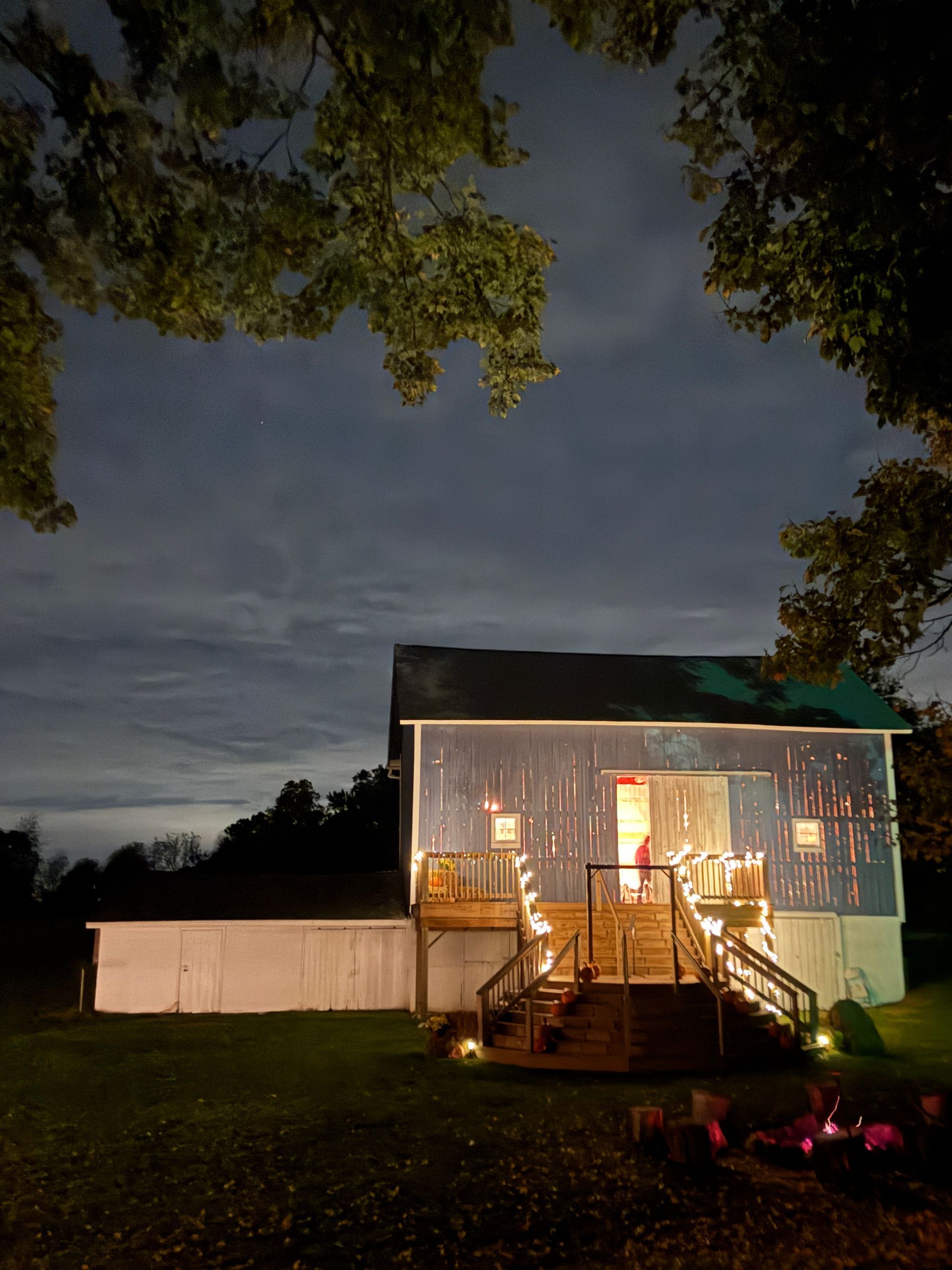 Blue wedding and event venue barn with a lit deck at night, framed by dark trees and a cloudy sky.