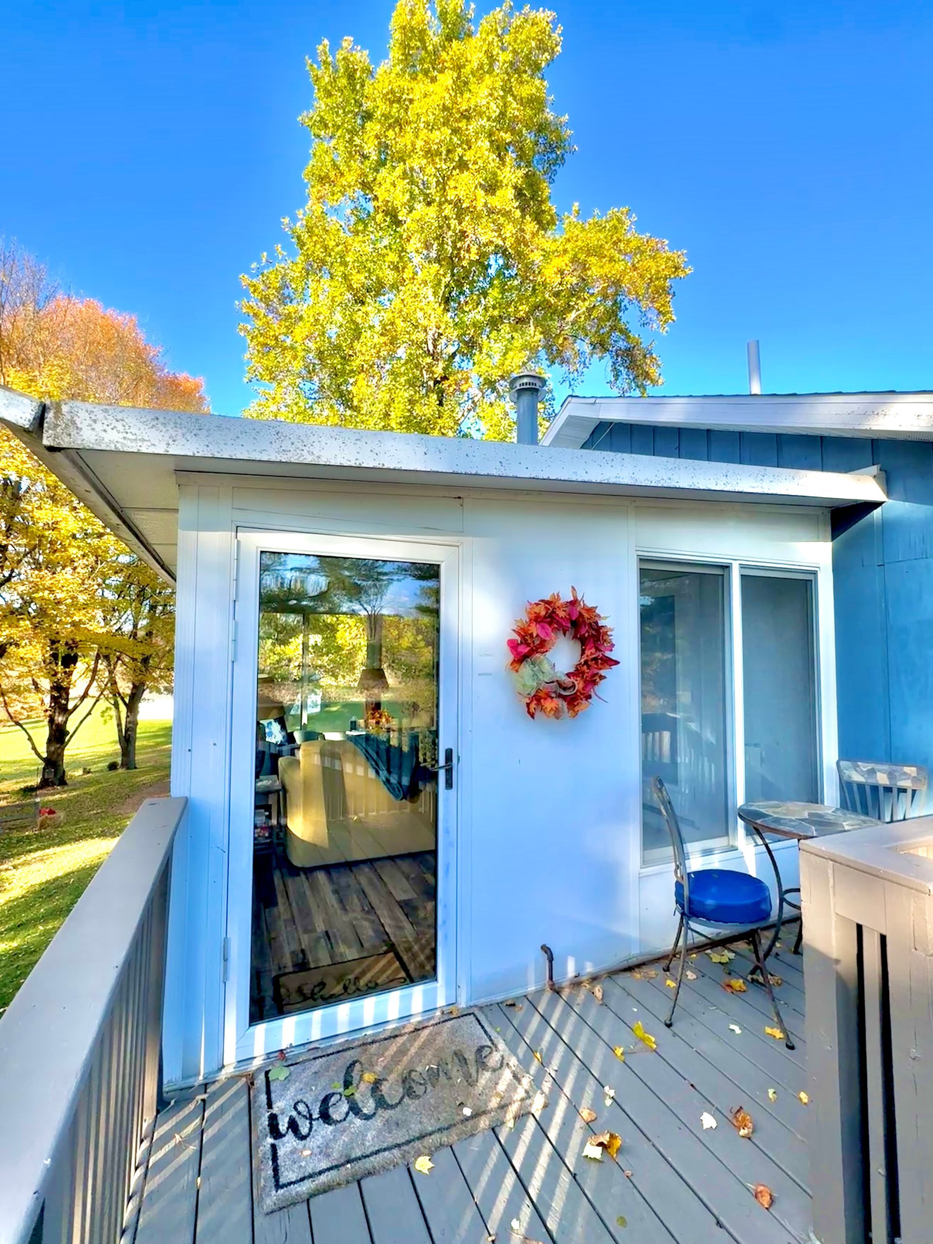 White-walled porch with fall wreath, glass door, and yellow-leafed tree under a blue sky.
