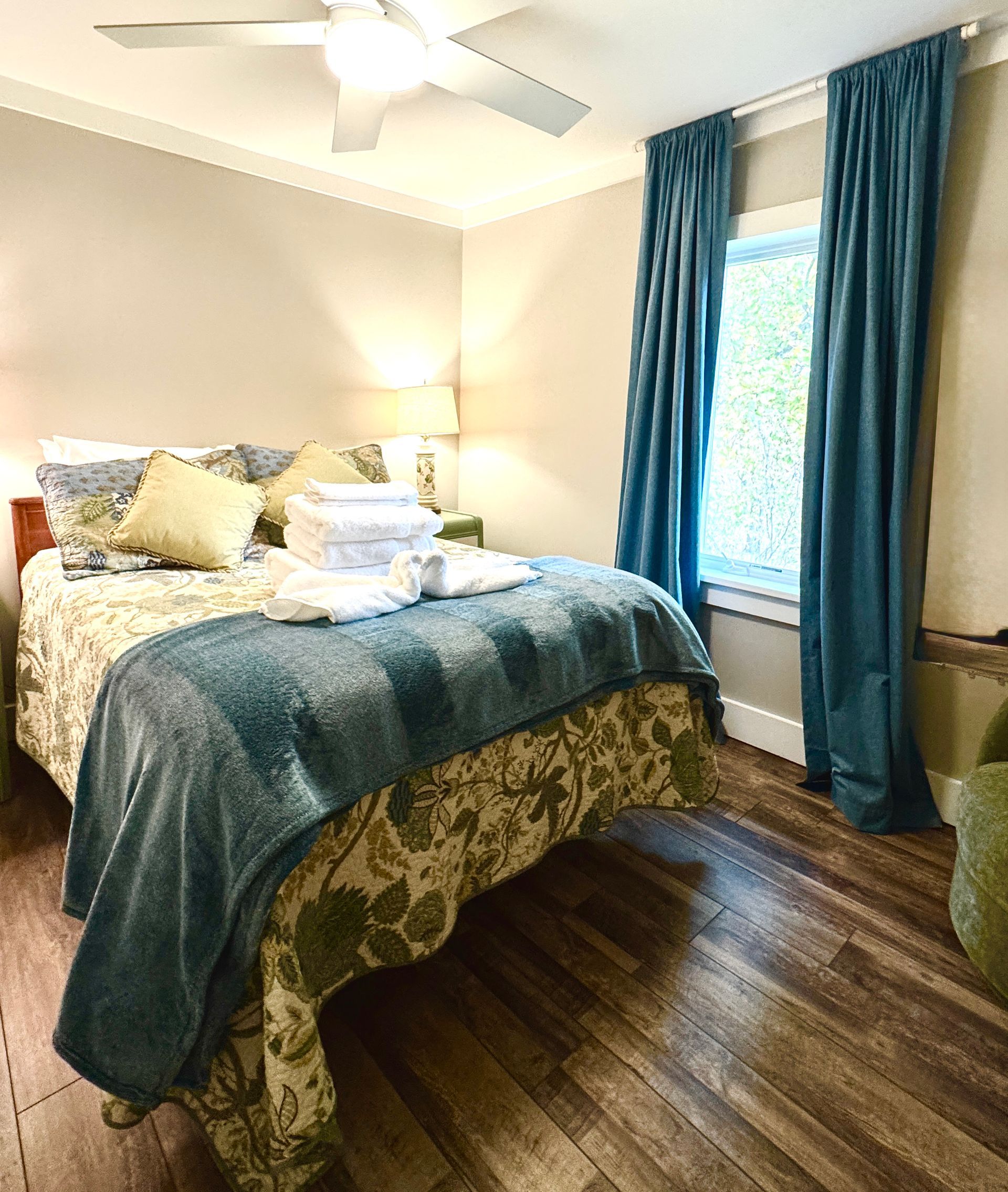 Bedroom with double bed, blue blanket, green patterned bedspread, blue black out curtains, and wood flooring.