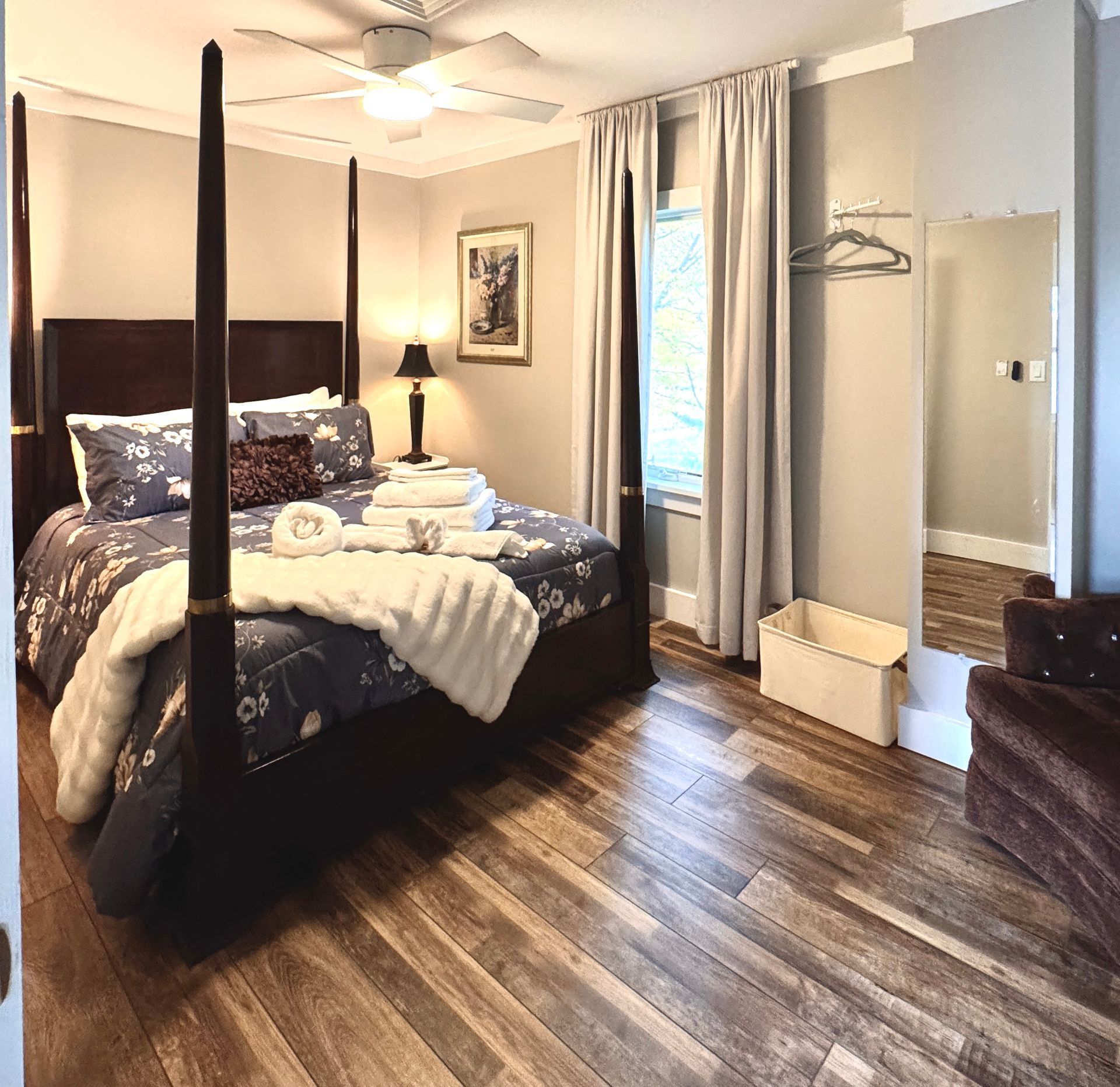 Bedroom with dark wood four-poster bed, floral bedding, black out curtains, hardwood floors, and a tall mirror.