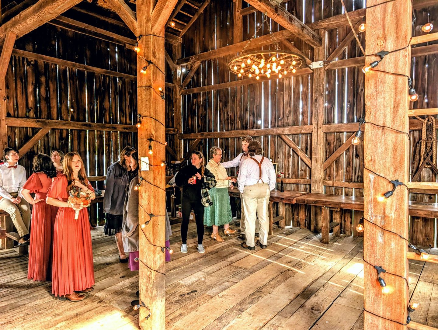 Wedding guests gathered inside a rustic barn. String lights illuminate the wooden beams and a central wagon wheel chandelier.