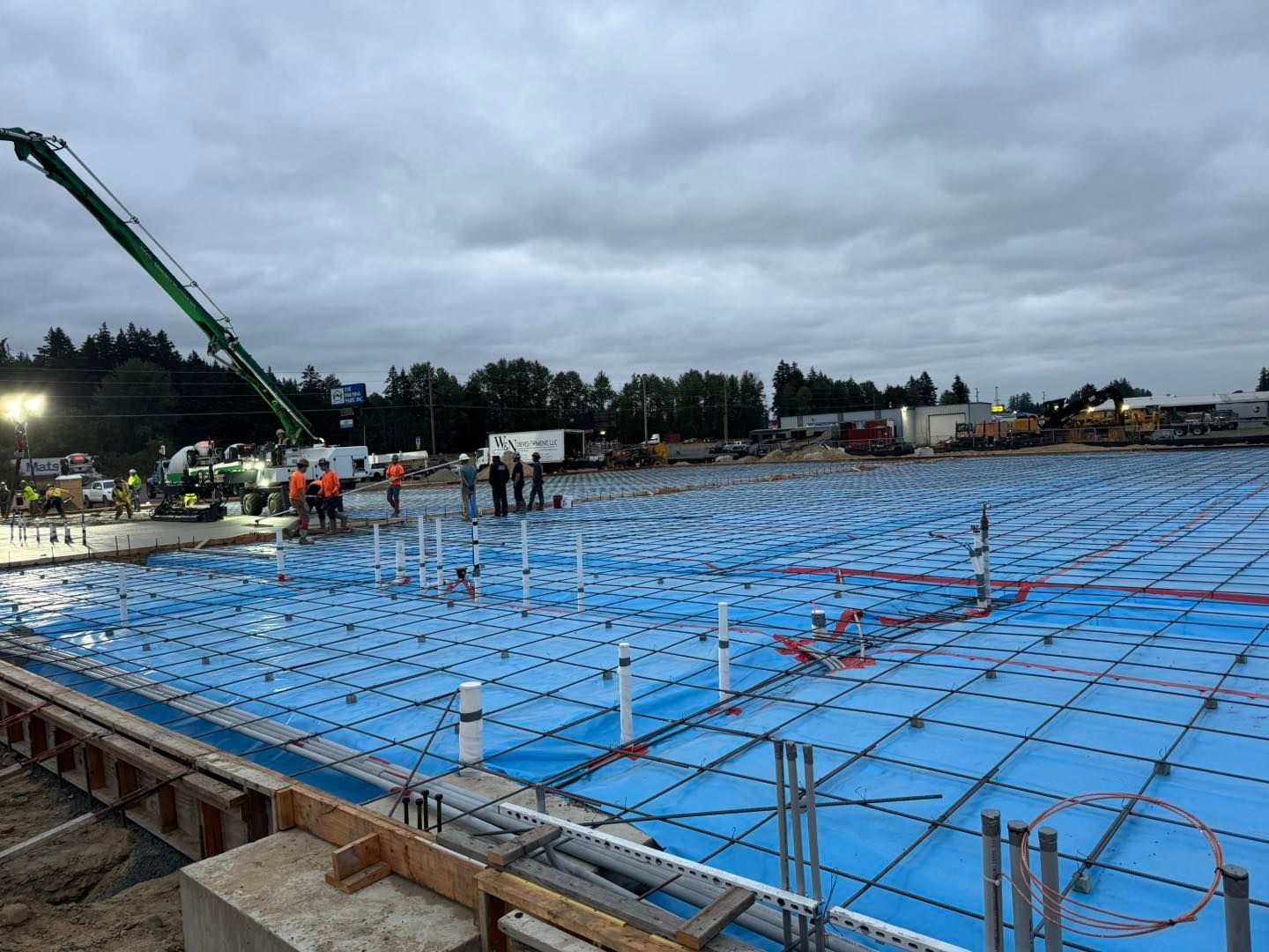 Construction site with blue waterproofing membrane over a large foundation slab and workers in the distance