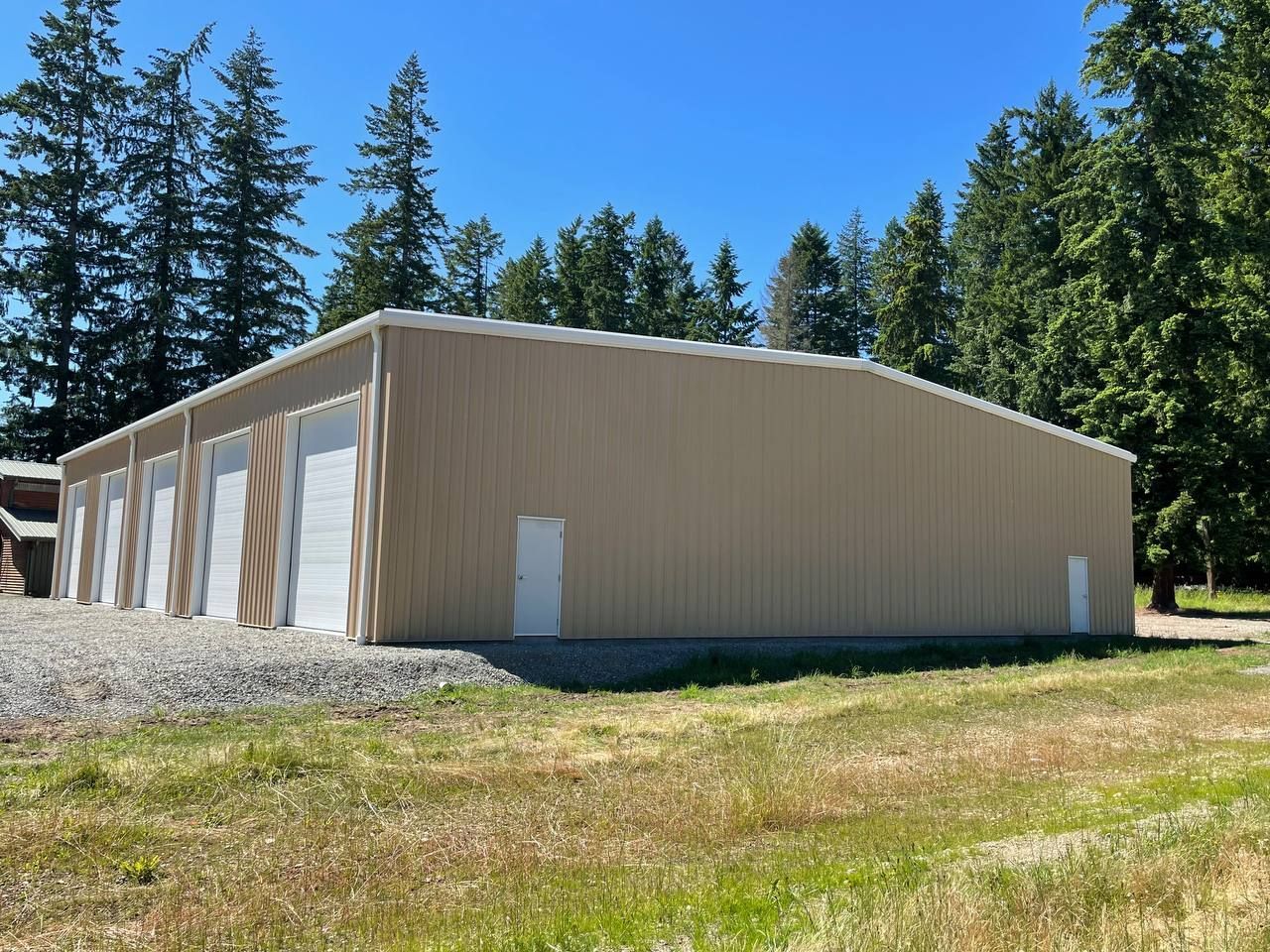 Beige metal warehouse with white doors on a grassy lot, surrounded by pine trees under a clear blue sky