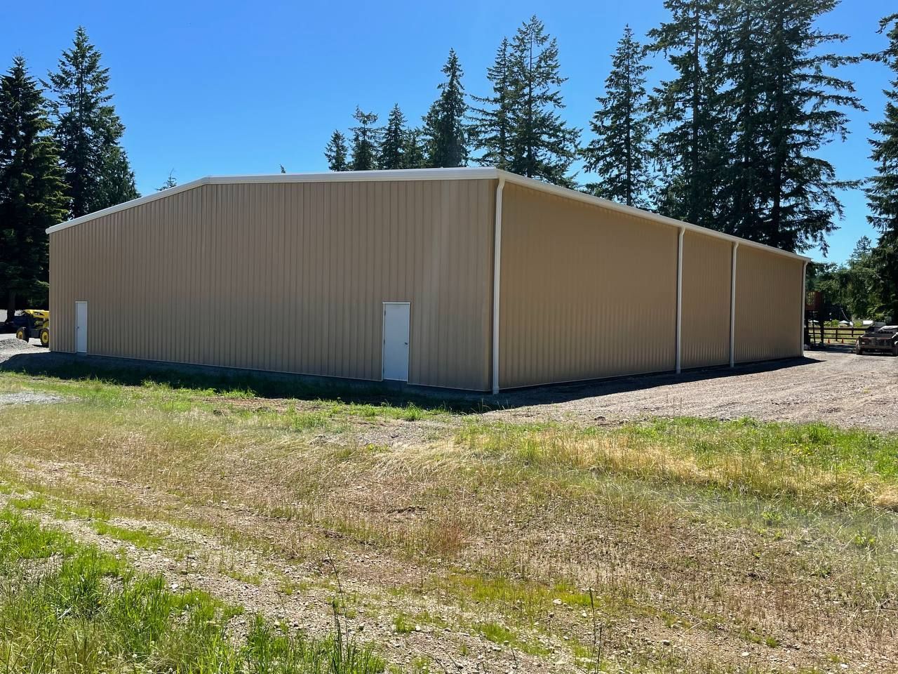 Beige metal storage building on a grassy lot with tall evergreen trees under a clear blue sky