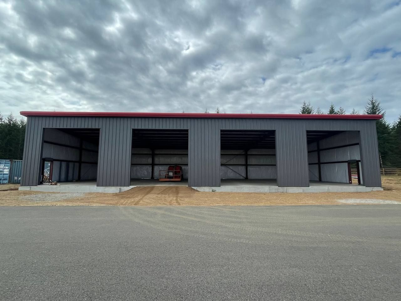 Large gray metal barn with open bays and a red roof beside a paved lot under a cloudy sky