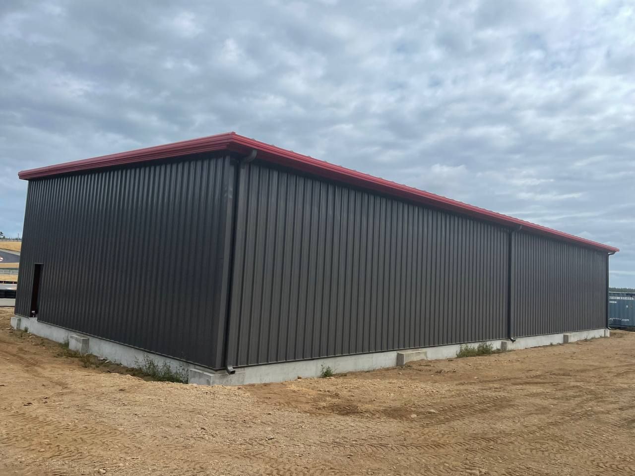 Large gray metal shed with a red roof on a dirt lot under a cloudy sky
