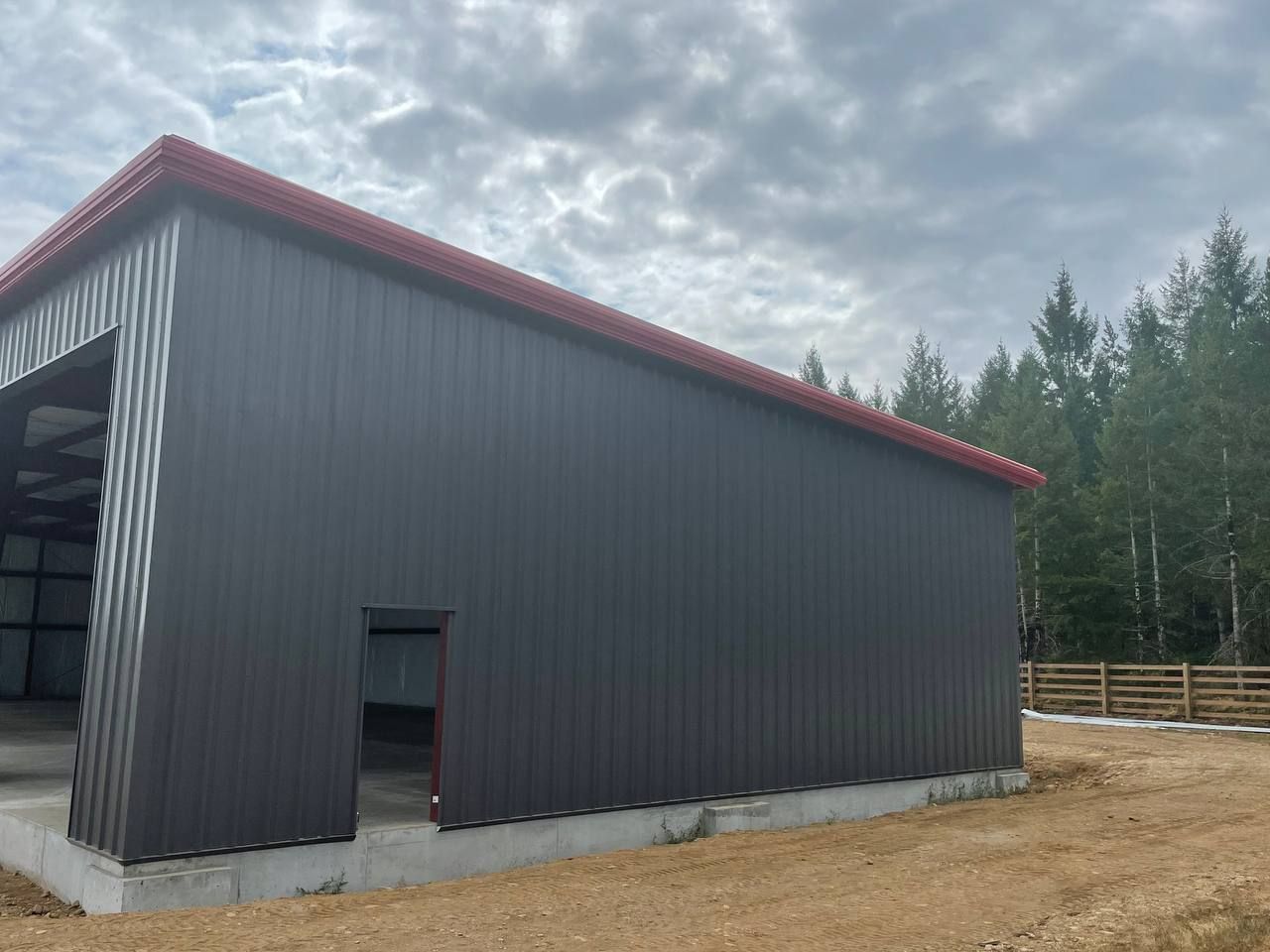 Black metal barn with red roof trim on a dirt lot under cloudy sky, viewed from the side.