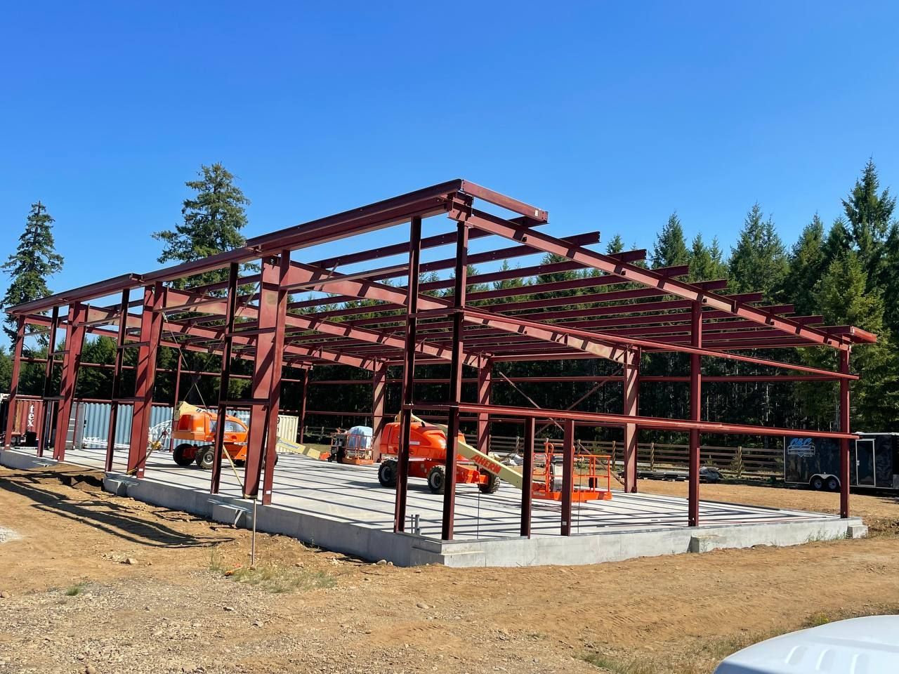 Steel frame of a building under construction on a concrete slab, with orange equipment and a clear blue sky.