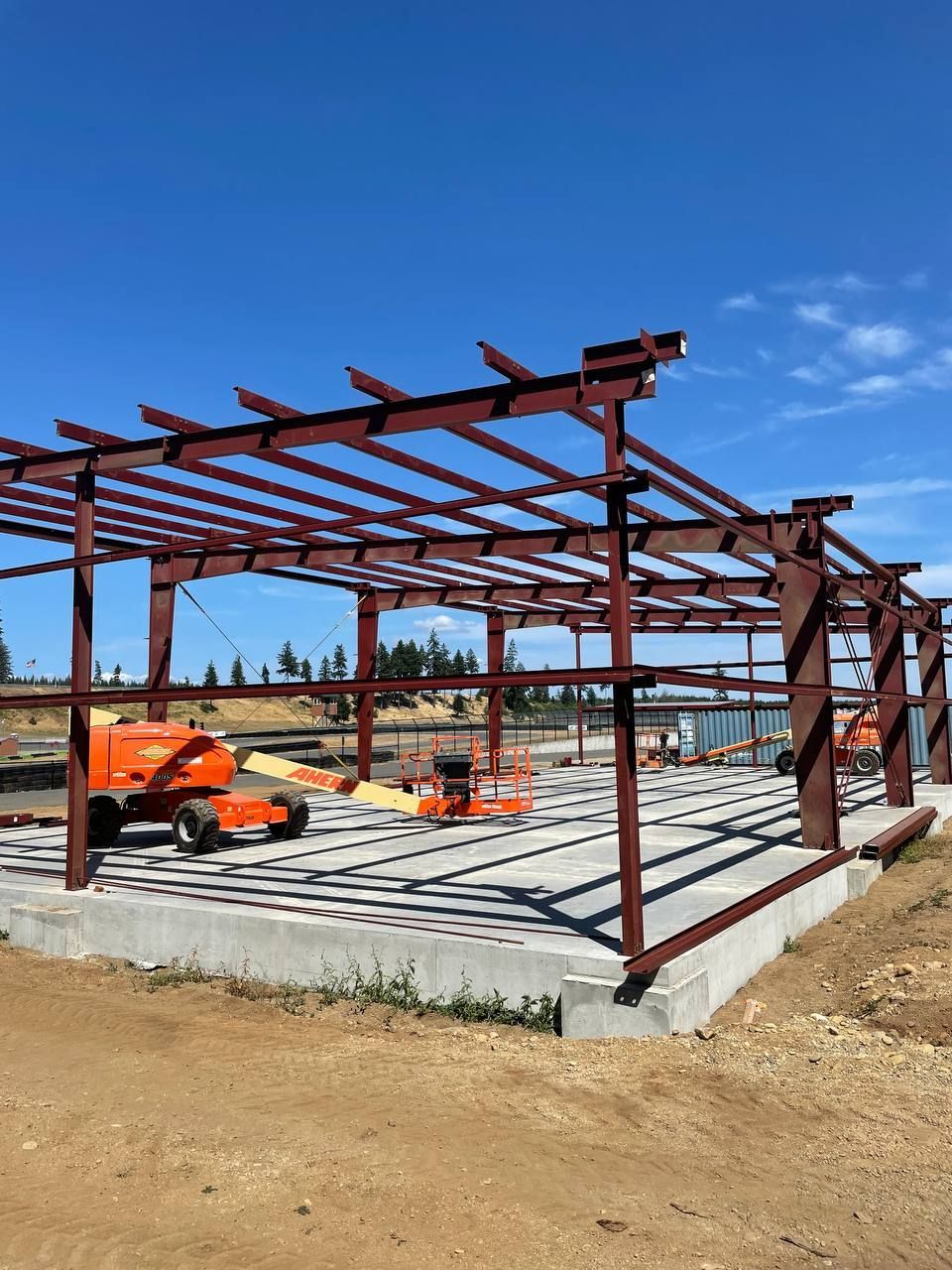 Steel building frame under construction with concrete foundation and orange lift equipment on site