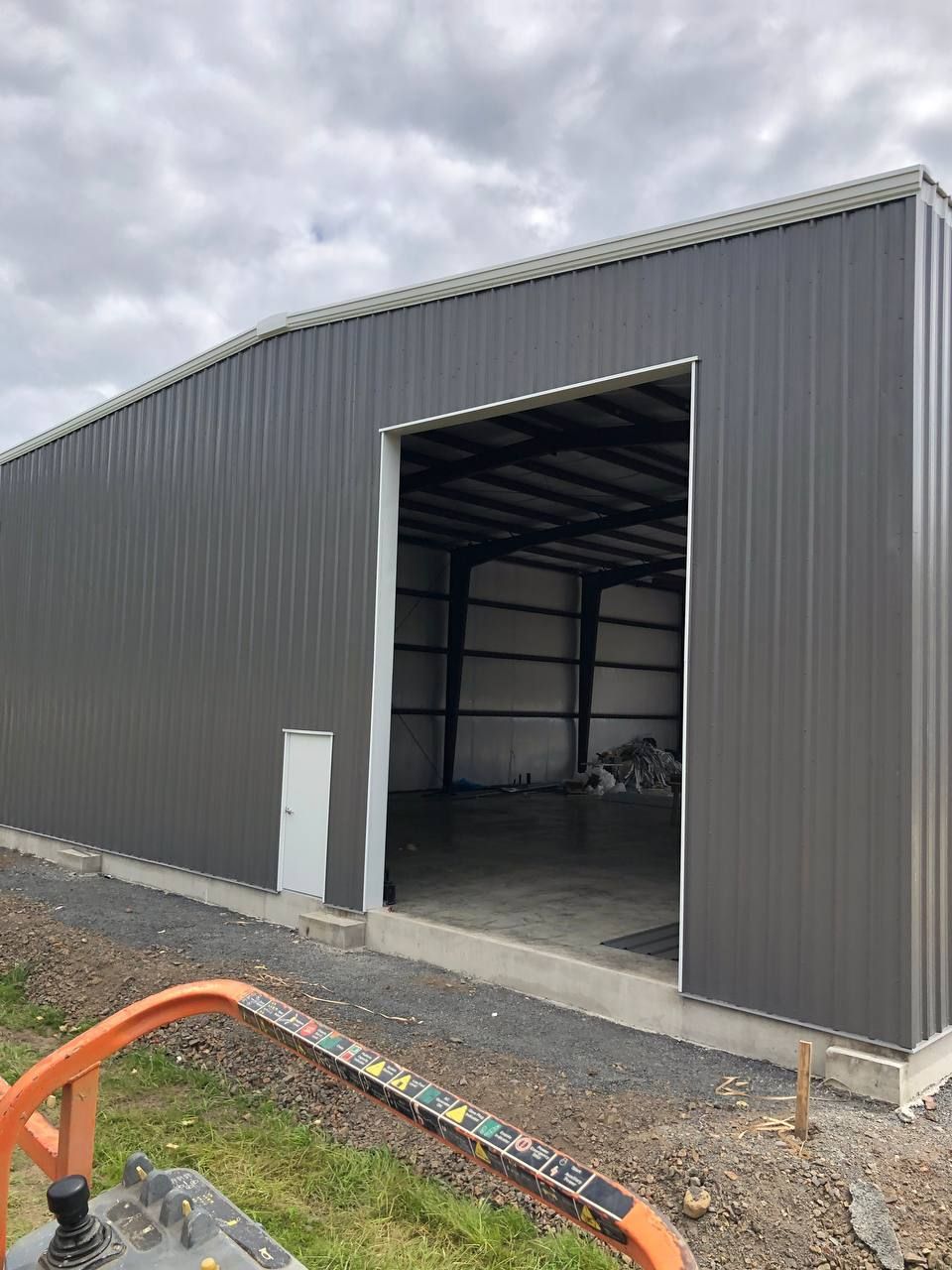 Large gray metal warehouse with open loading bay and gravel foreground under cloudy sky