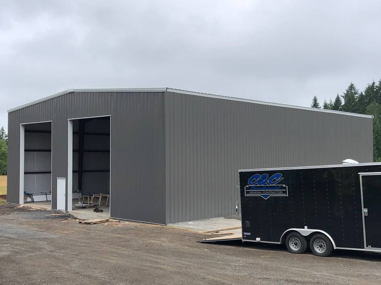 Large gray metal building under construction with an open doorway and a black trailer in front