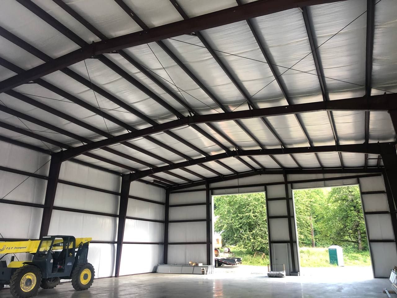 Interior of a metal warehouse with a yellow forklift and large open doors showing trees outside