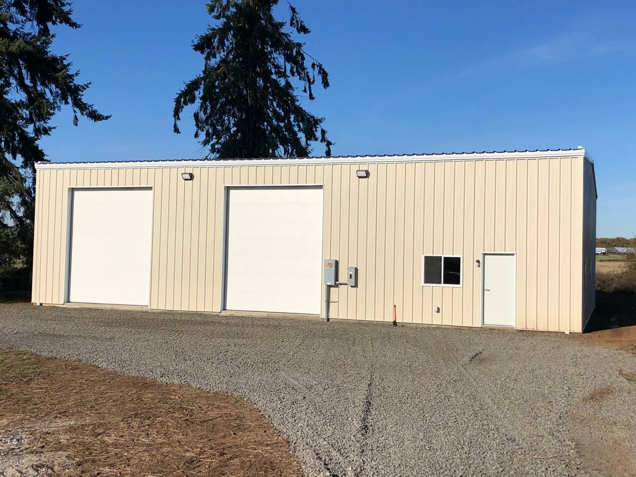 Beige metal storage building with two large garage doors and a side entry on a gravel lot under a blue sky