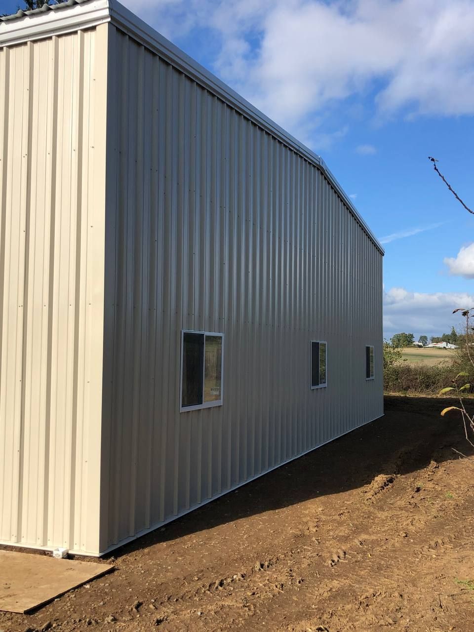 Metal-sided building under construction on a dirt lot, with small windows and a blue sky.