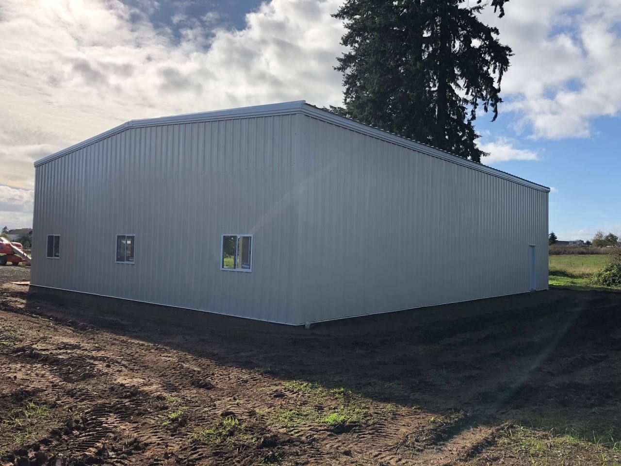 White metal warehouse building on a dirt lot with a tree behind it and fields in the background
