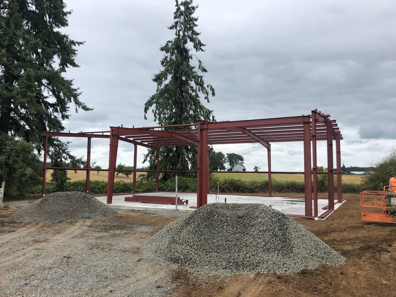 Red steel frame under construction on a gravel site with conifer trees and cloudy sky in the background