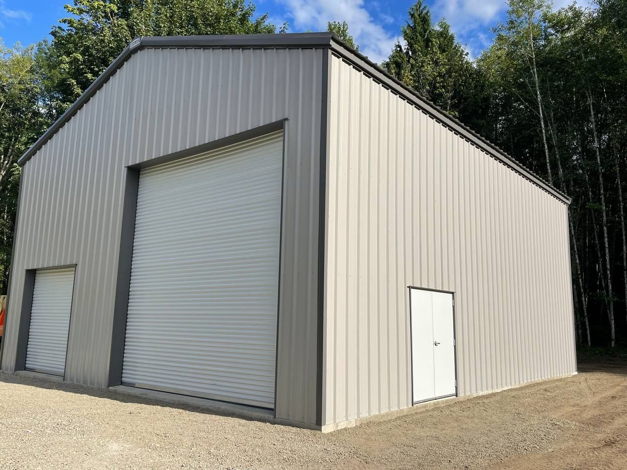 Large gray metal storage building with roll-up doors and a white side door on a gravel lot