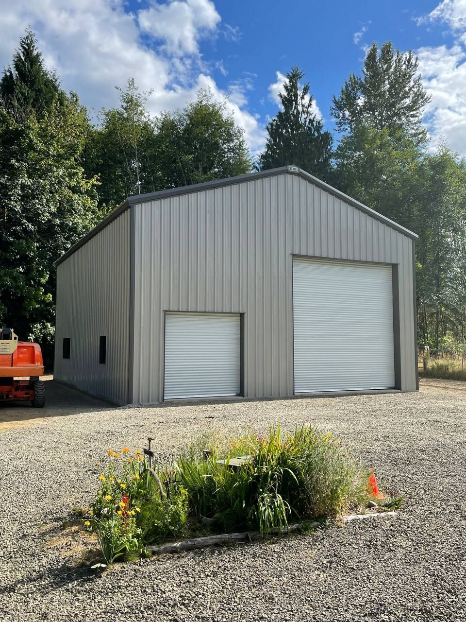 Gray metal storage building with two roll-up doors on a gravel lot, trees and blue sky behind it
