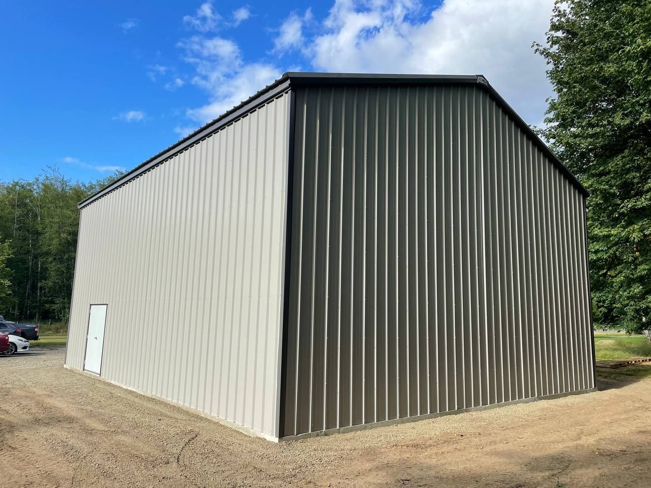 Large beige metal barn with a dark roof on a dirt lot under a blue sky.