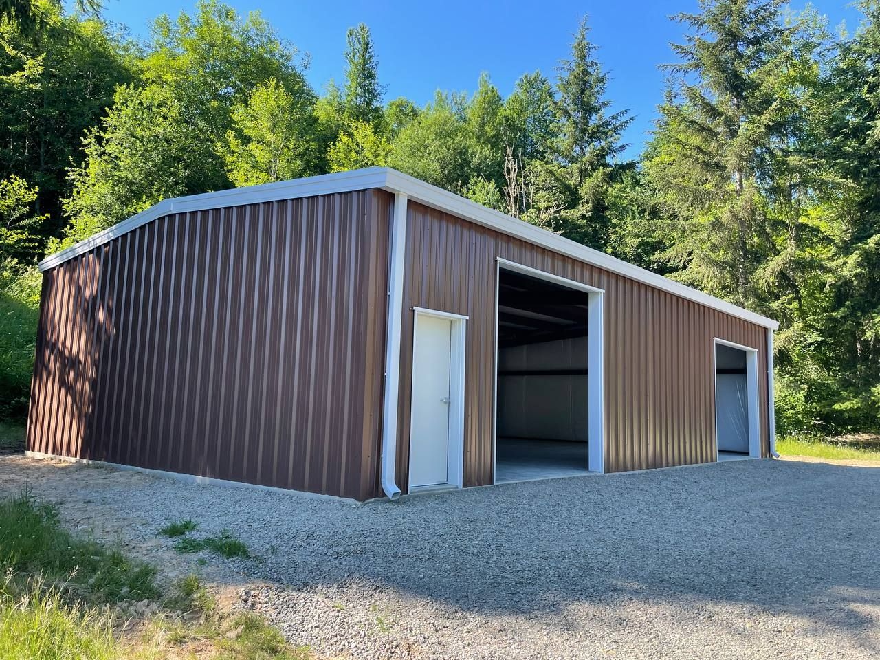 Brown metal shed with open garage door on a gravel lot, surrounded by trees and a blue sky