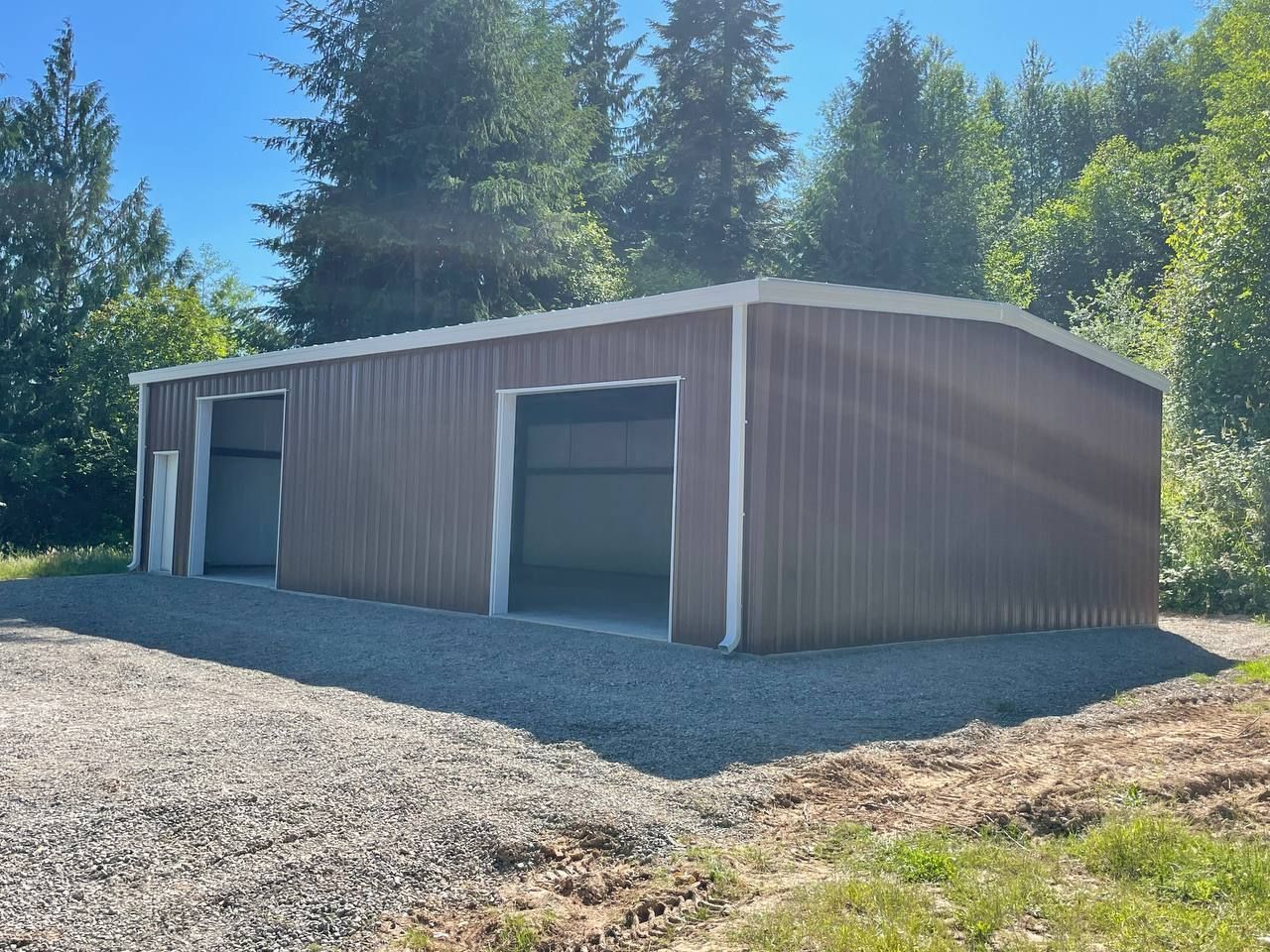 Brown metal storage building with open garage doors on a gravel lot surrounded by trees
