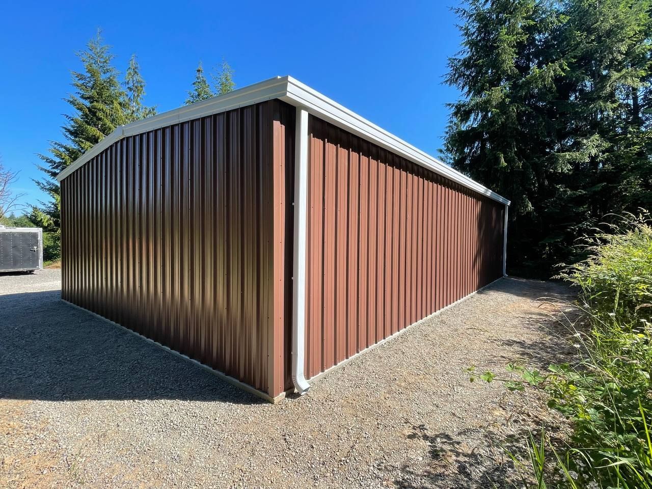 Brown metal storage shed on a gravel lot, viewed from the corner against a blue sky.
