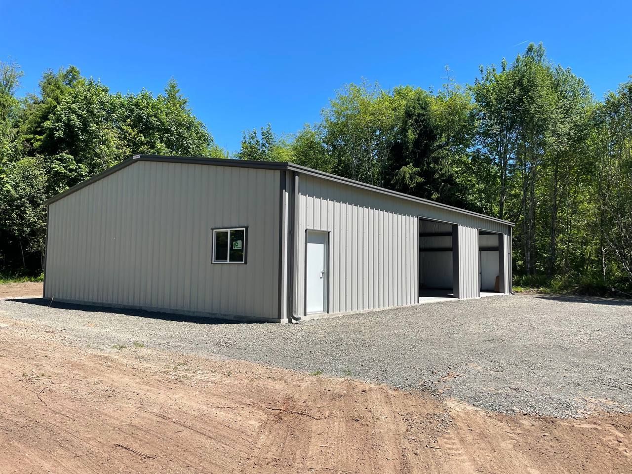 Gray metal storage building with open bay doors on a gravel lot, surrounded by trees under a blue sky