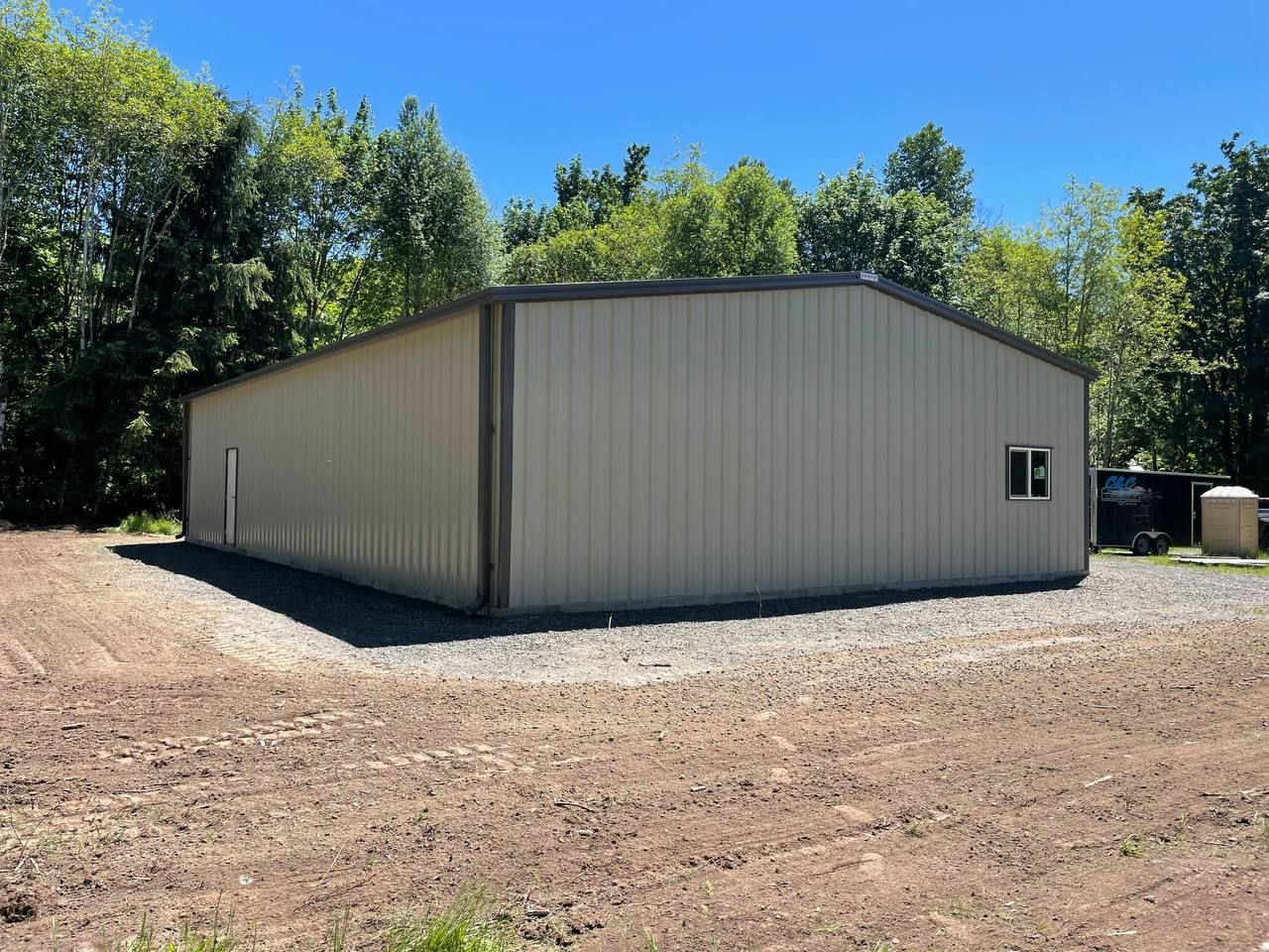 Large beige metal storage building on a dirt lot surrounded by trees under a blue sky